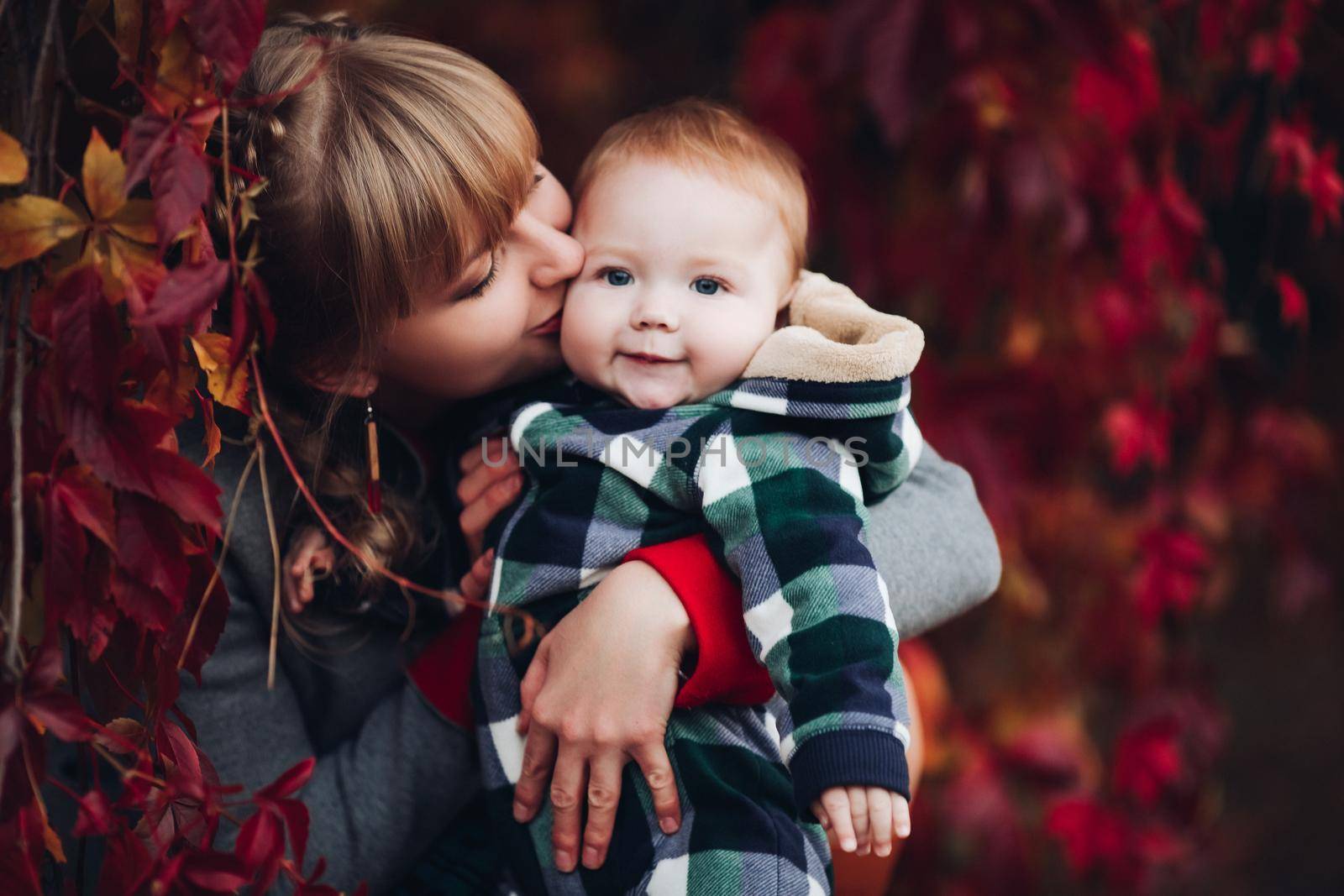 Professional portrait of attractive young woman with fair hair in braid embracing her little baby girl in plaid warm overall while standing in beautiful bright tree with red leaves. They are smiling at camera surrounded by vivid foliage in autumnal park.