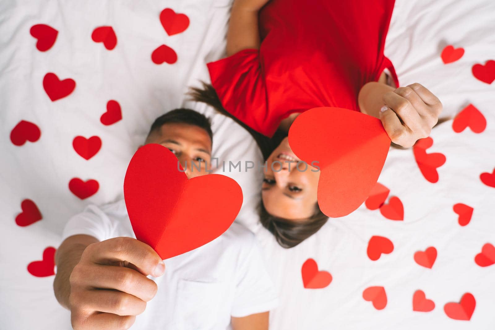 Top view of young attractive couple in love lying on the bed and showing red paper hearts. Happy valentine's day concept. High quality photo