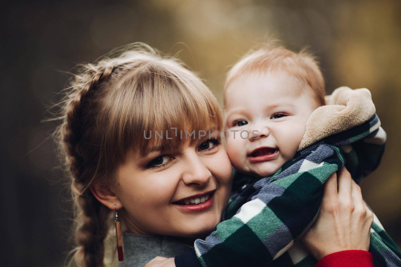 Professional portrait of attractive young woman with fair hair in braid embracing her little baby girl in plaid warm overall while standing in beautiful bright tree with red leaves. They are smiling at camera surrounded by vivid foliage in autumnal park.