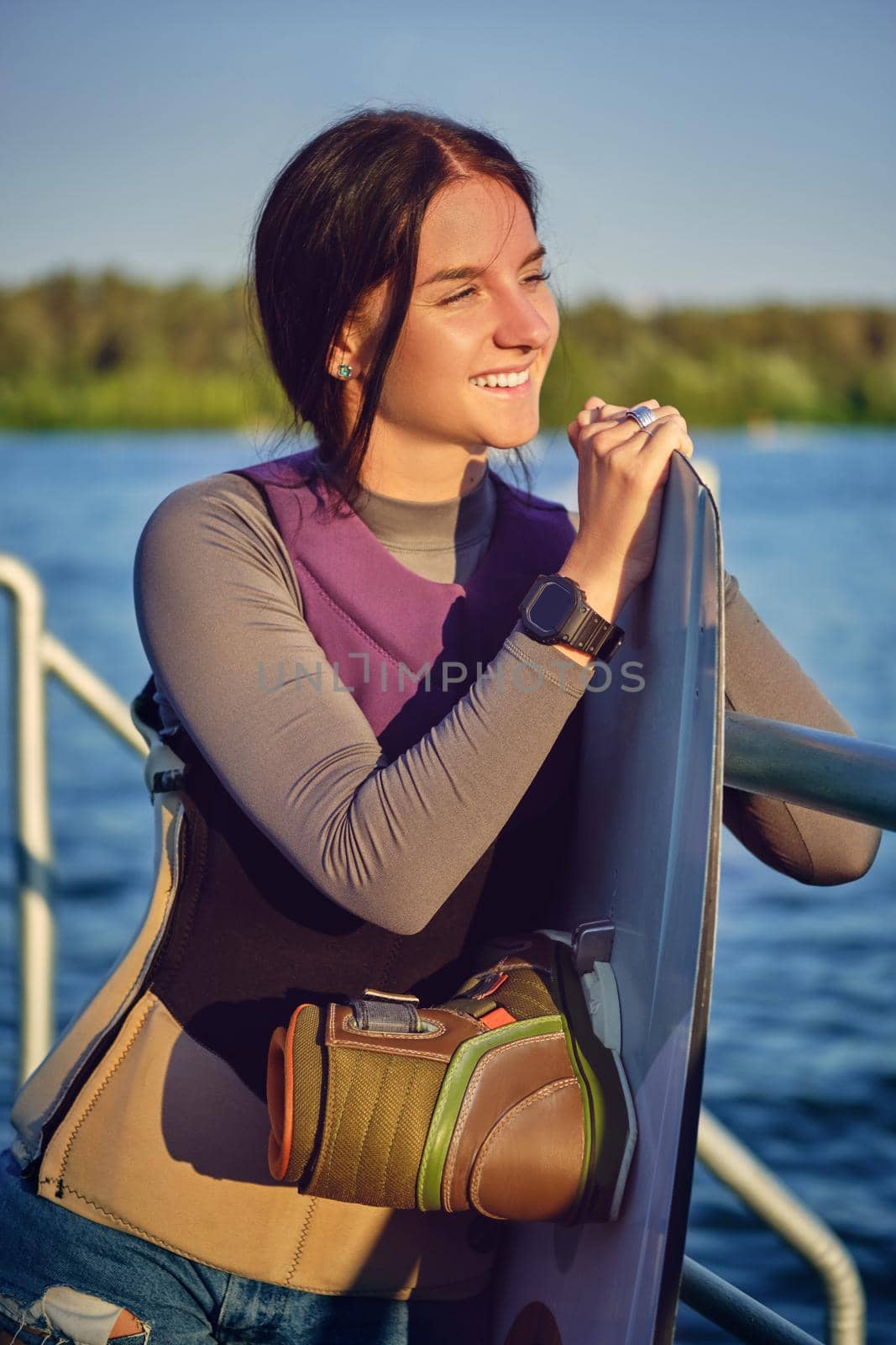 Alluring brunette woman in a gray turtleneck, special sports vest and black watch is looking away and smiling while posing with her wakeboard and standing on a pier of the riverside. Sport and recreation concept. Close-up shot.