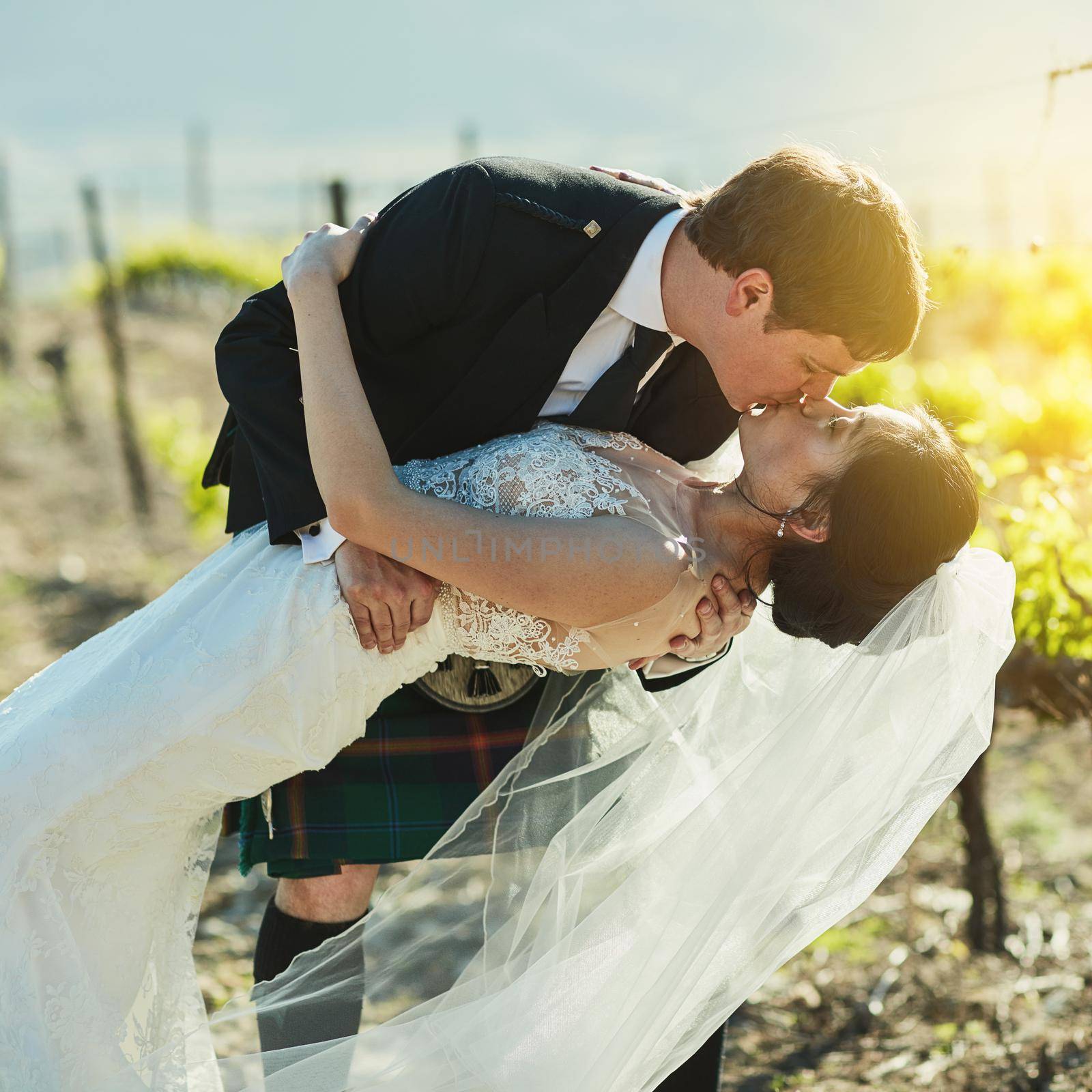 What a stunning kiss. Shot of a cheerful bride and groom sharing a kiss together outside next to vineyards during the day. by YuriArcurs