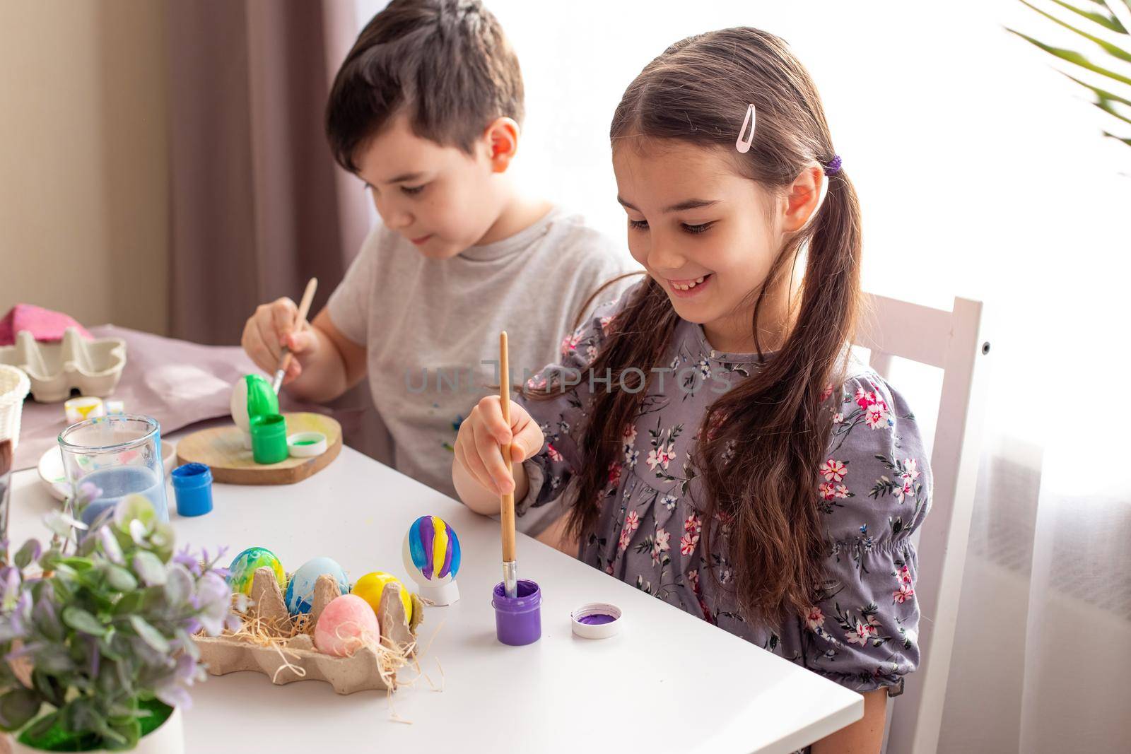 A smiling little girl and boy sits at a white table near the window, prepares for the Easter holiday, paints eggs