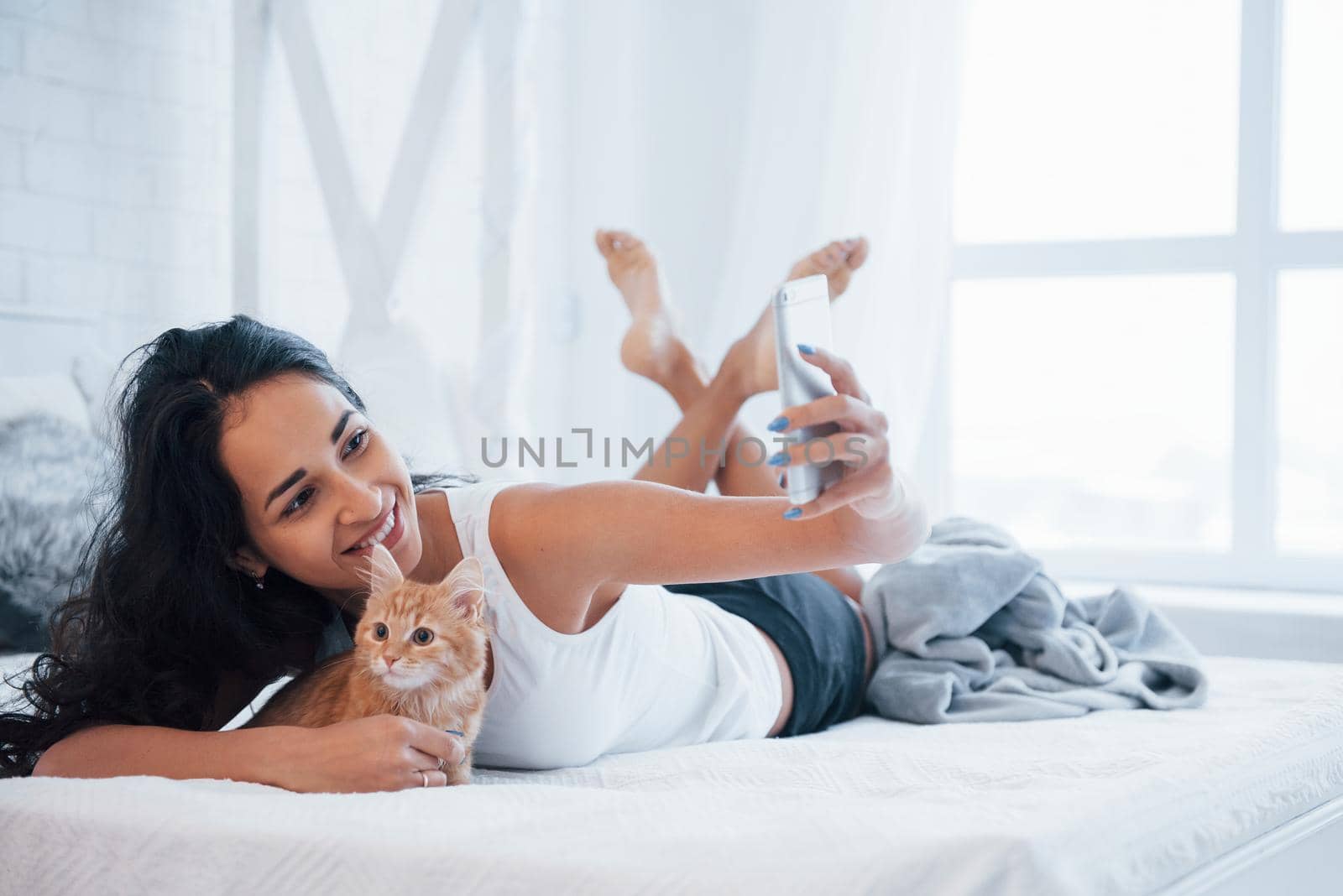 Taking selfie. Attractive blonde resting on the white bed with her cute kitten by Standret