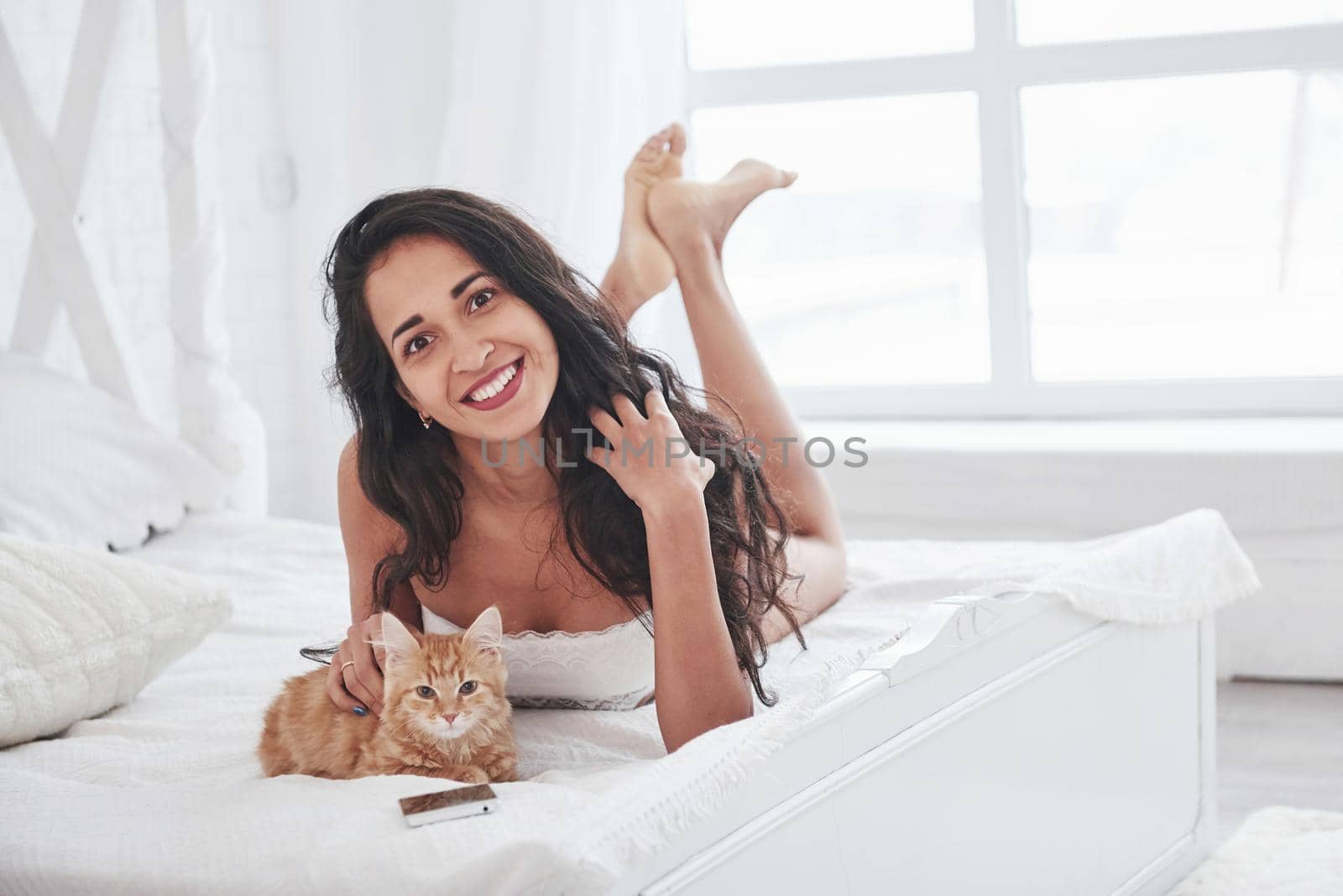 Such a sincere smile. Attractive blonde resting on the white bed with her cute kitten.