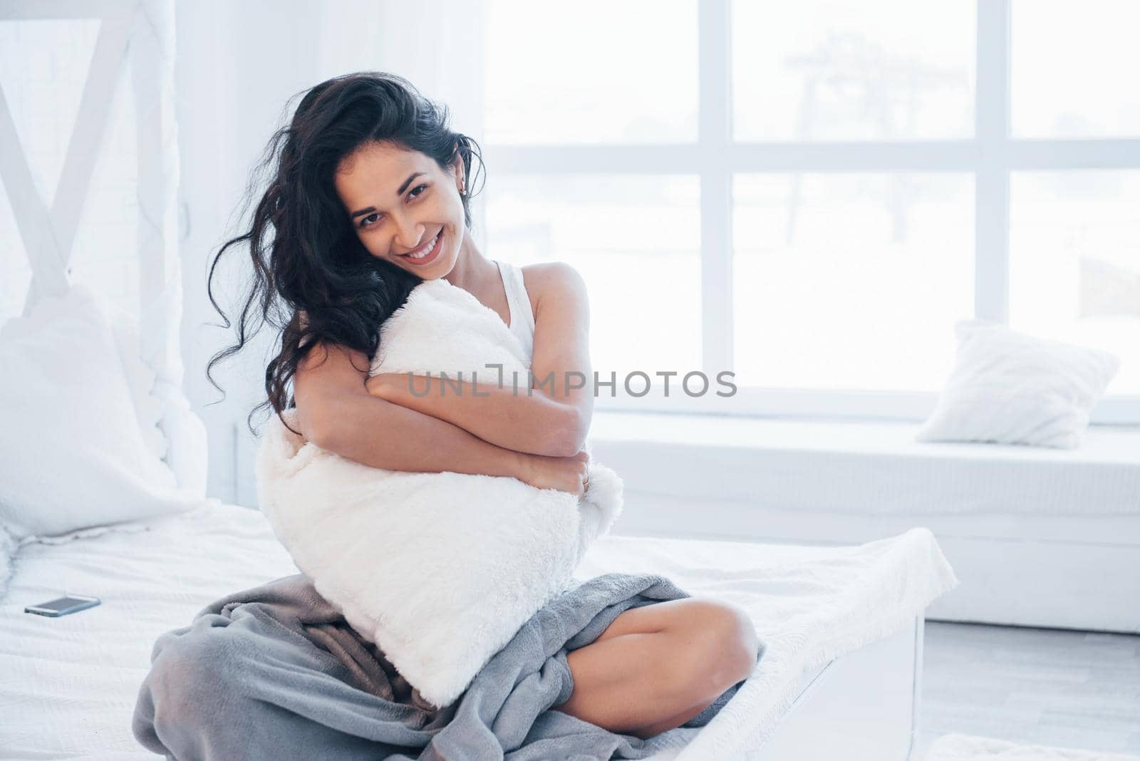 Hugging a pillow. Brunette having a rest on the white couch in her bedroom at daytime.