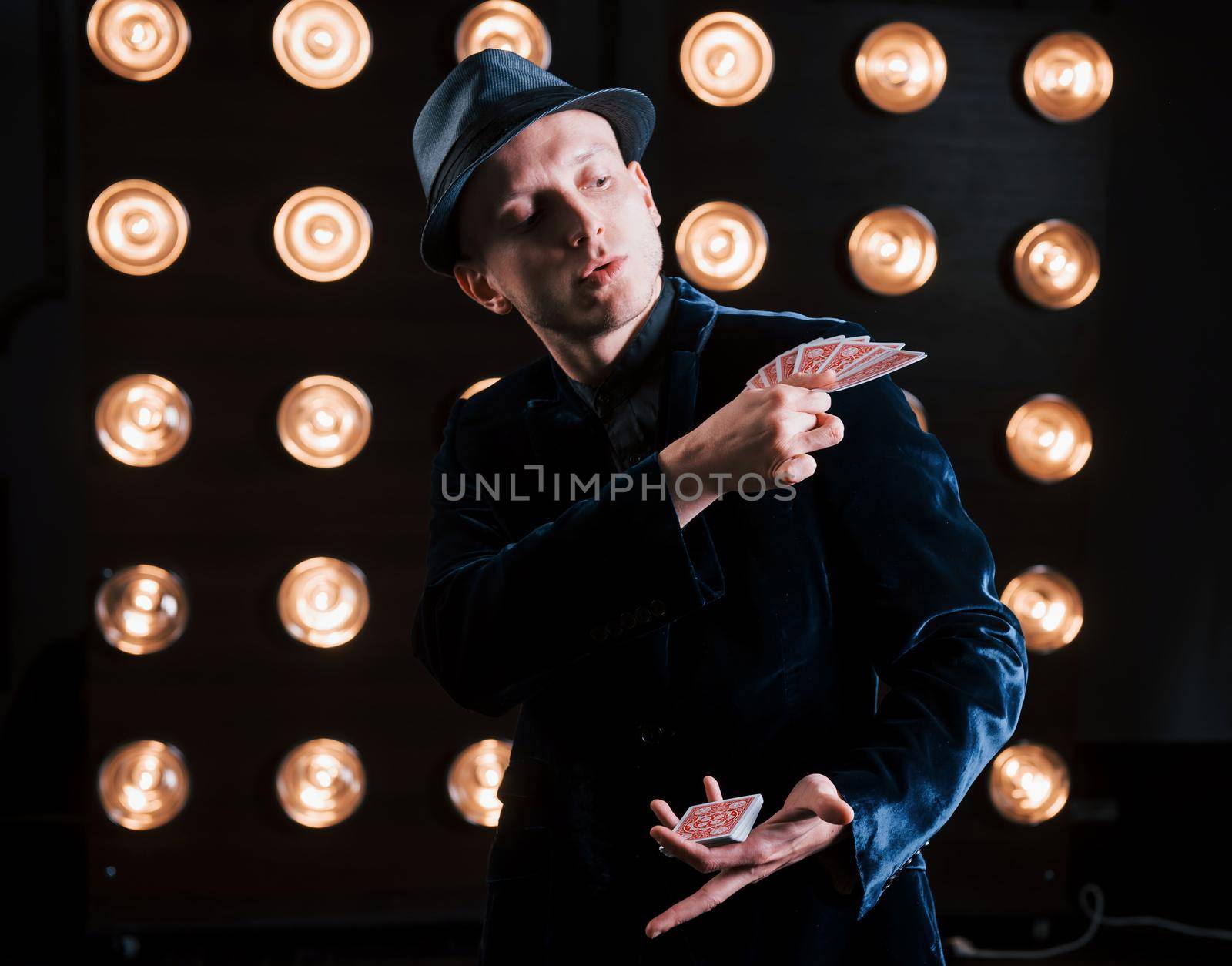 Showing professional tricks. Magician in black suit and with playing cards standing in the room with special lighting at backstage.