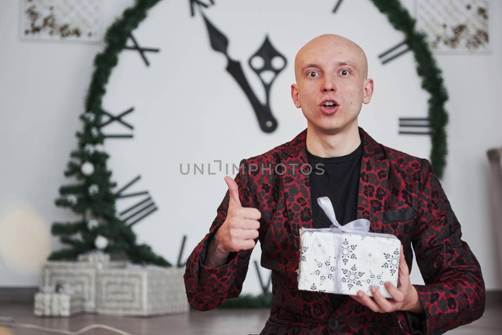 Sjowing thumb up. Man with big clock behind him sitting with New year gift boxes in holiday clothings.