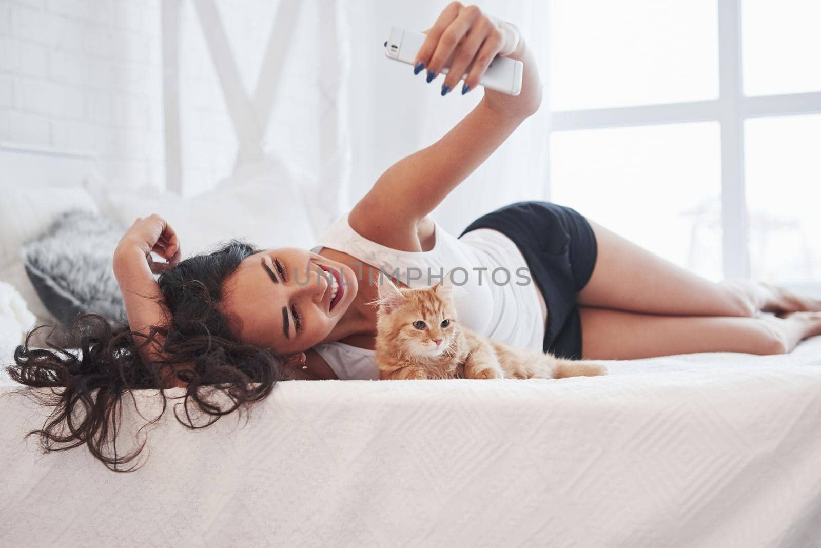 Relaxed day wih phone in hands. Attractive blonde resting on the white bed with her cute kitten.