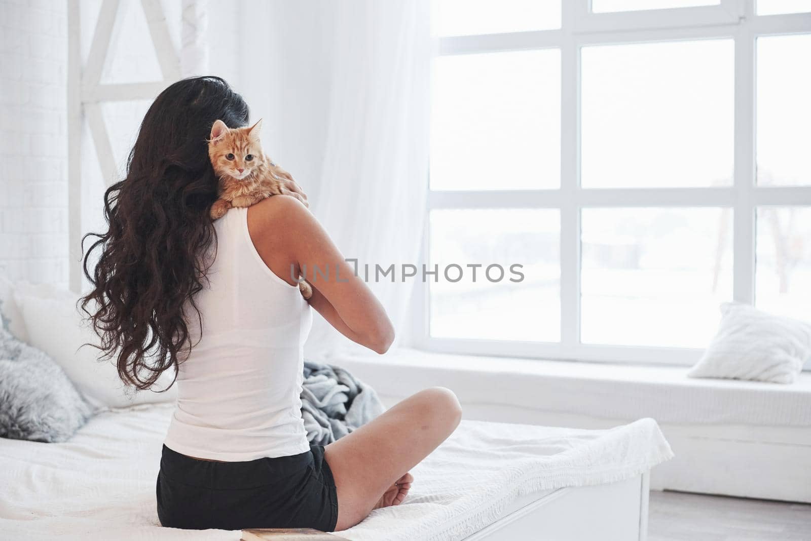 Pet sitting on the shoulder. Attractive blonde resting on the white bed with her cute kitten.