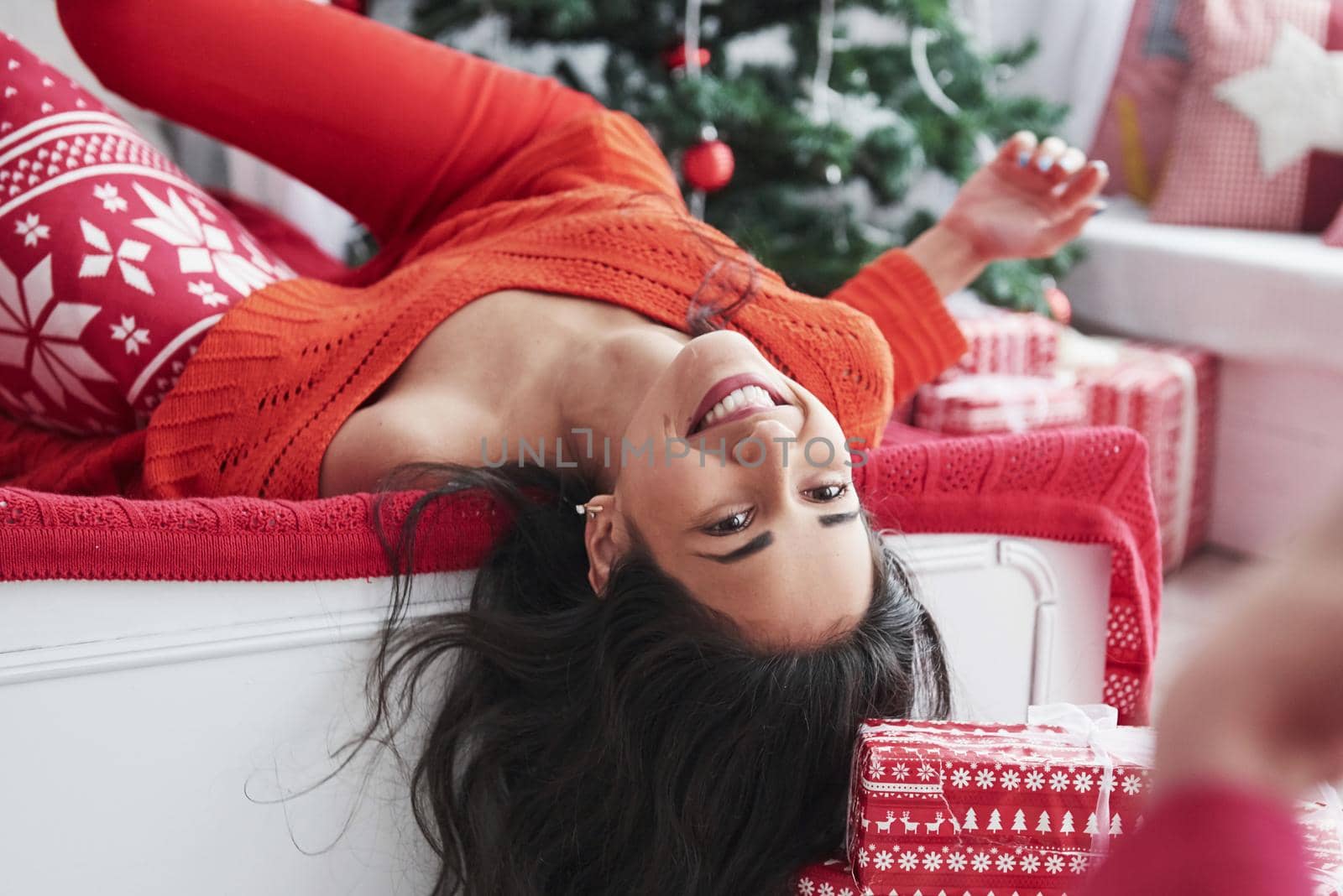 Upside down. Girl in red holiday clothes lying on the bed in the decorative bedroom with green tree behind.