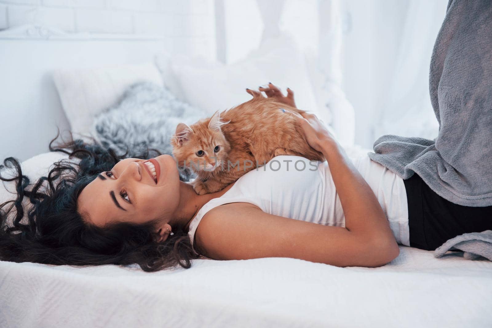 Looking up and smiling. Attractive blonde resting on the white bed with her cute kitten.