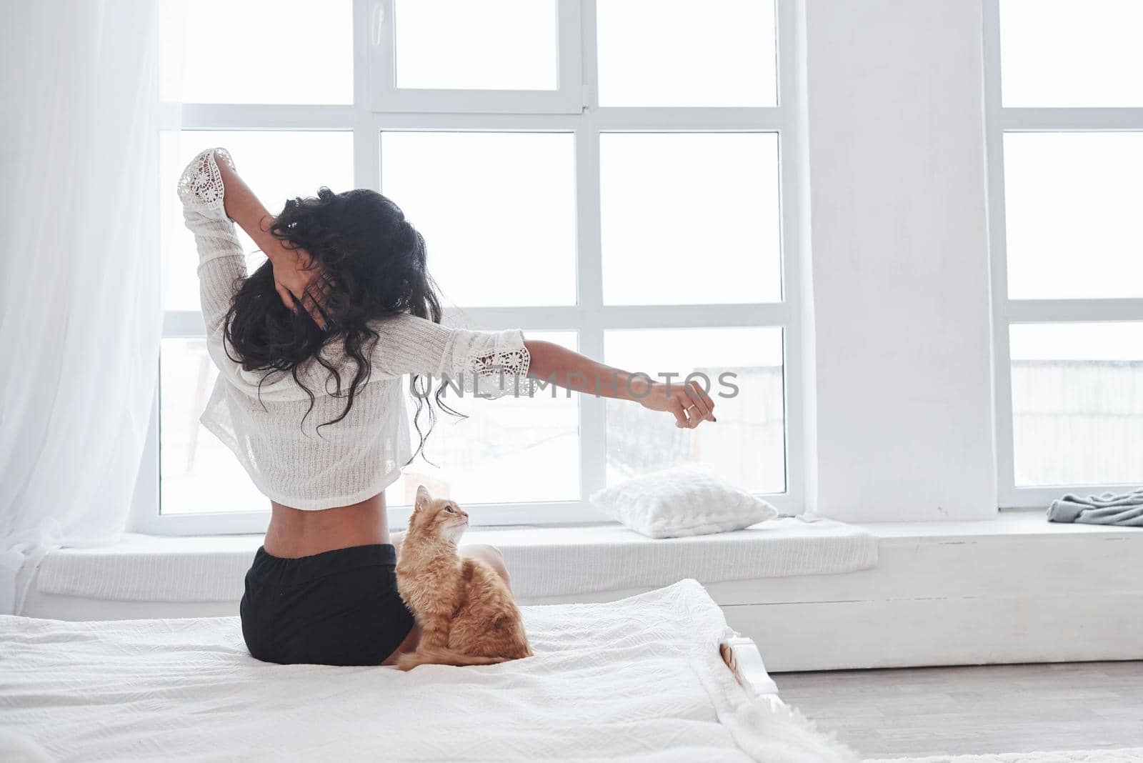 Spacious room. Attractive blonde resting on the white bed with her cute kitten.