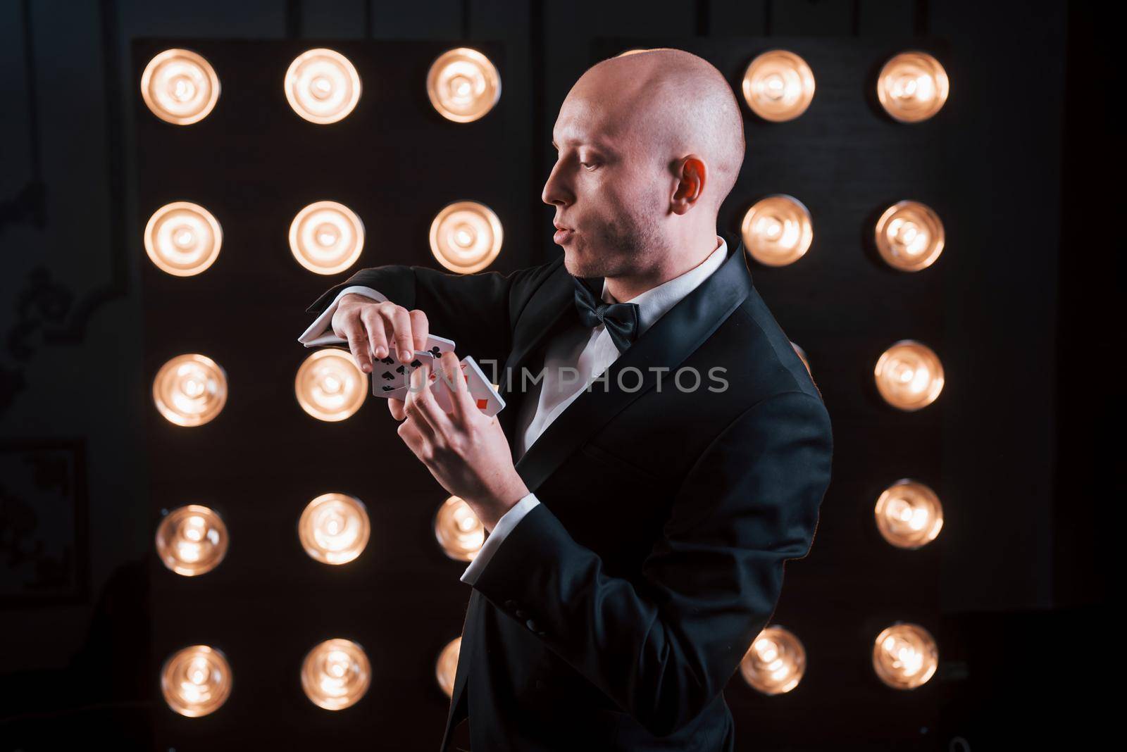 Concentrating at tricky move. Magician in black suit and with playing cards standing in the room with special lighting at backstage.