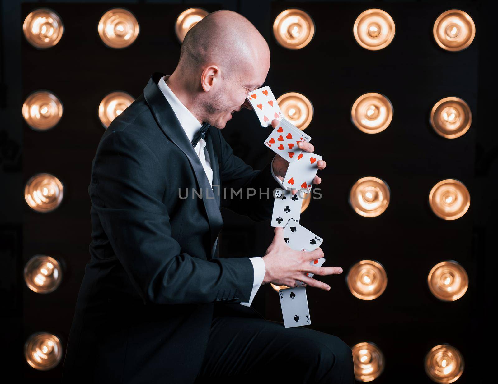 Makes it looks easy. Magician in black suit and with playing cards standing in the room with special lighting at backstage.