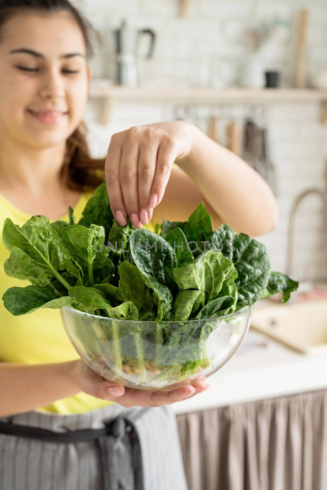 Preparing healthy foods. Healthy eating and dieting. Young caucasian brunette woman holding a bowl of fresh spinach in the kitchen