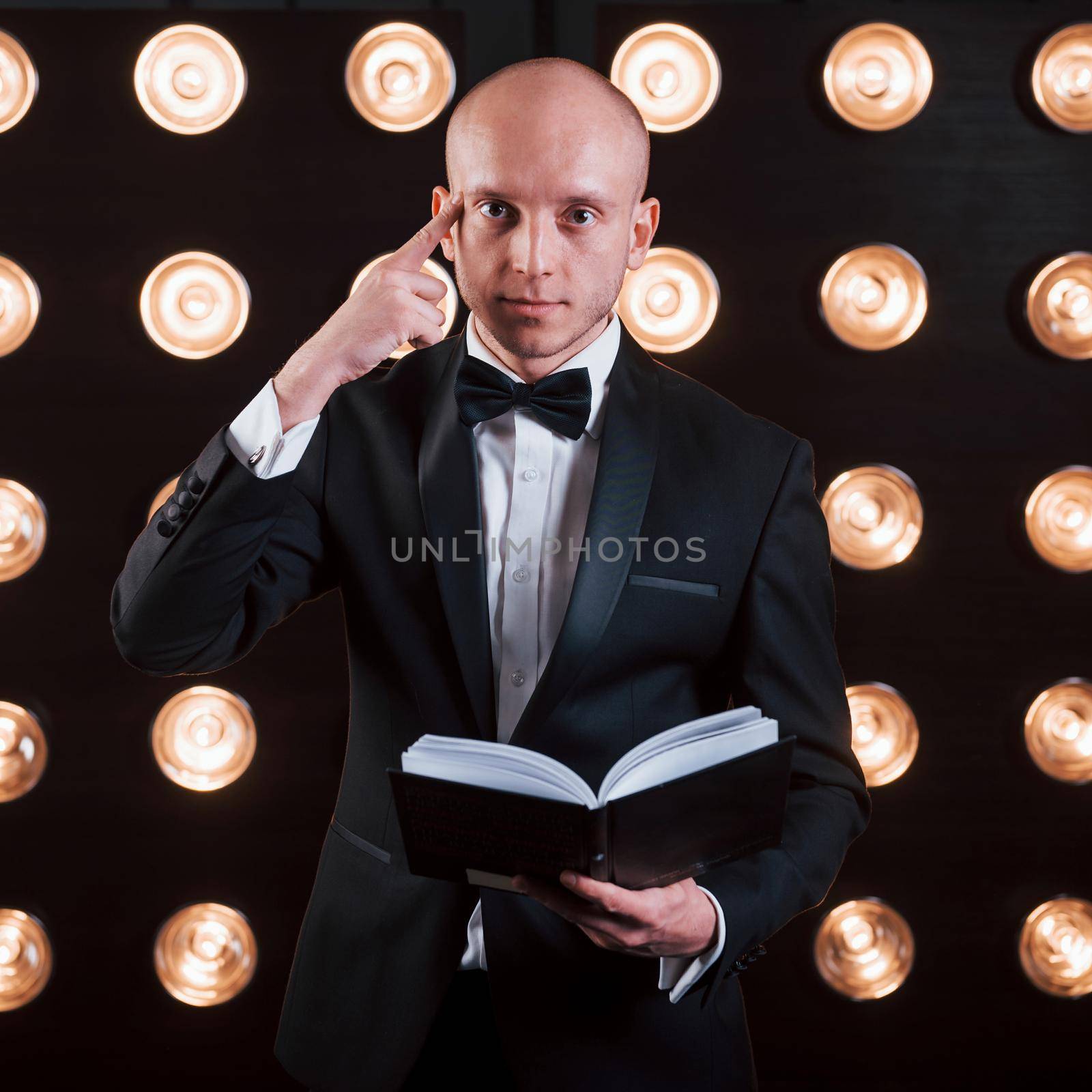 Reading your mind. Magician in black suit standing in the room with special lighting at backstage.