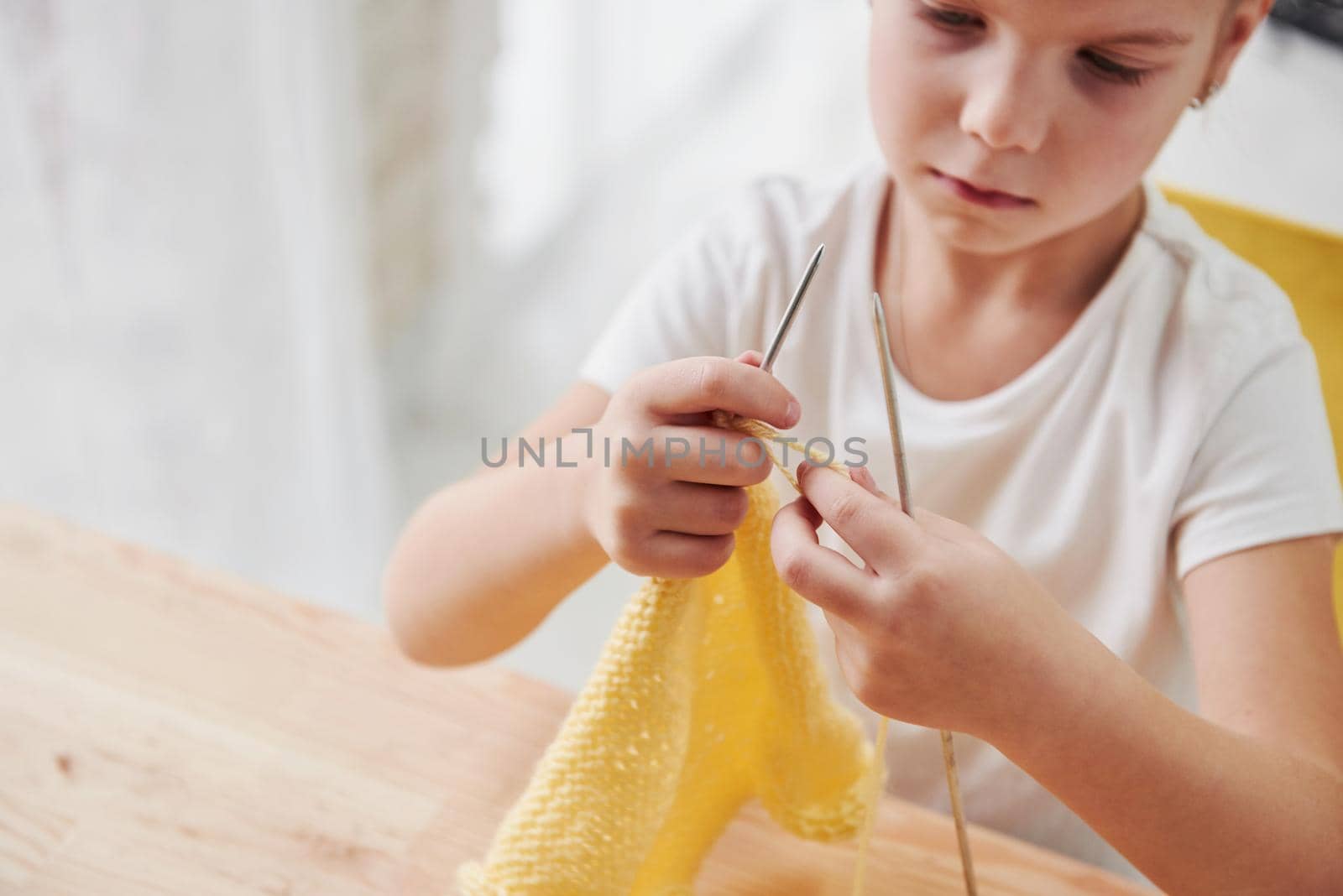Focused photo. Kid is knitting at home. Cute little girl sitting near the wooden table is learning some new stuff.