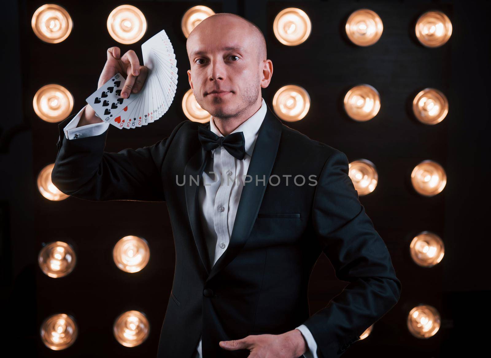 Concept of circus. Magician in black suit and with playing cards standing in the room with special lighting at backstage.