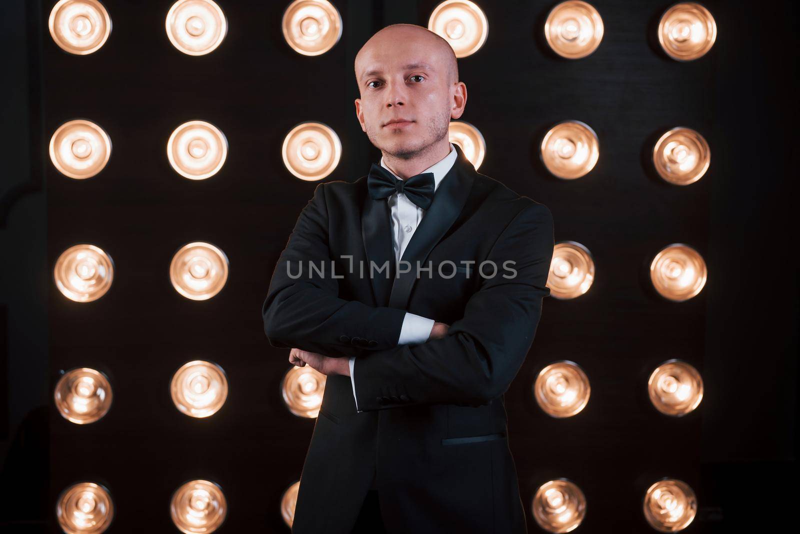 With arms crossed. Magician in black suit standing in the room with special lighting at backstage.