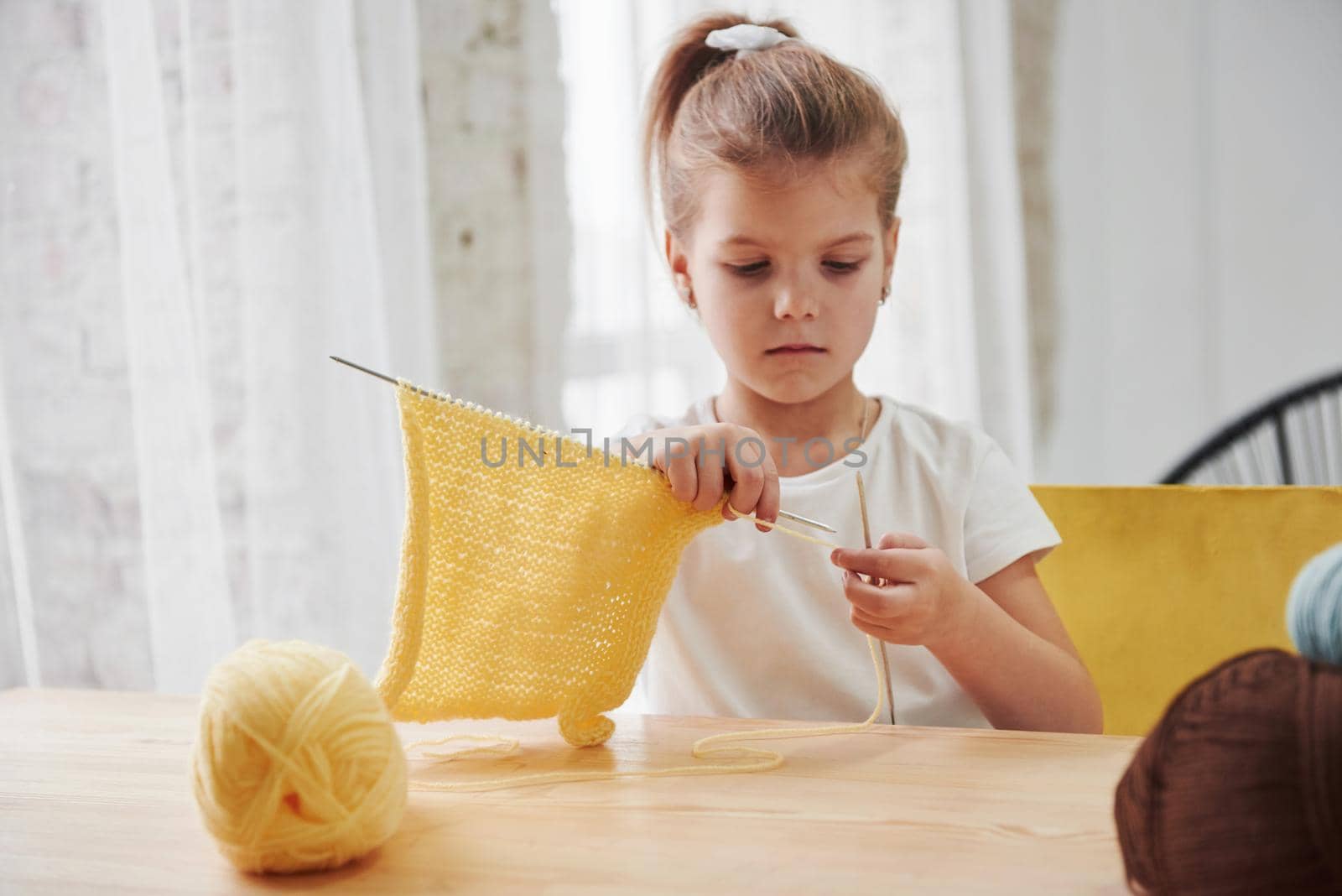 She's good at it. Kid is knitting at home. Cute little girl sitting near the wooden table is learning some new stuff.