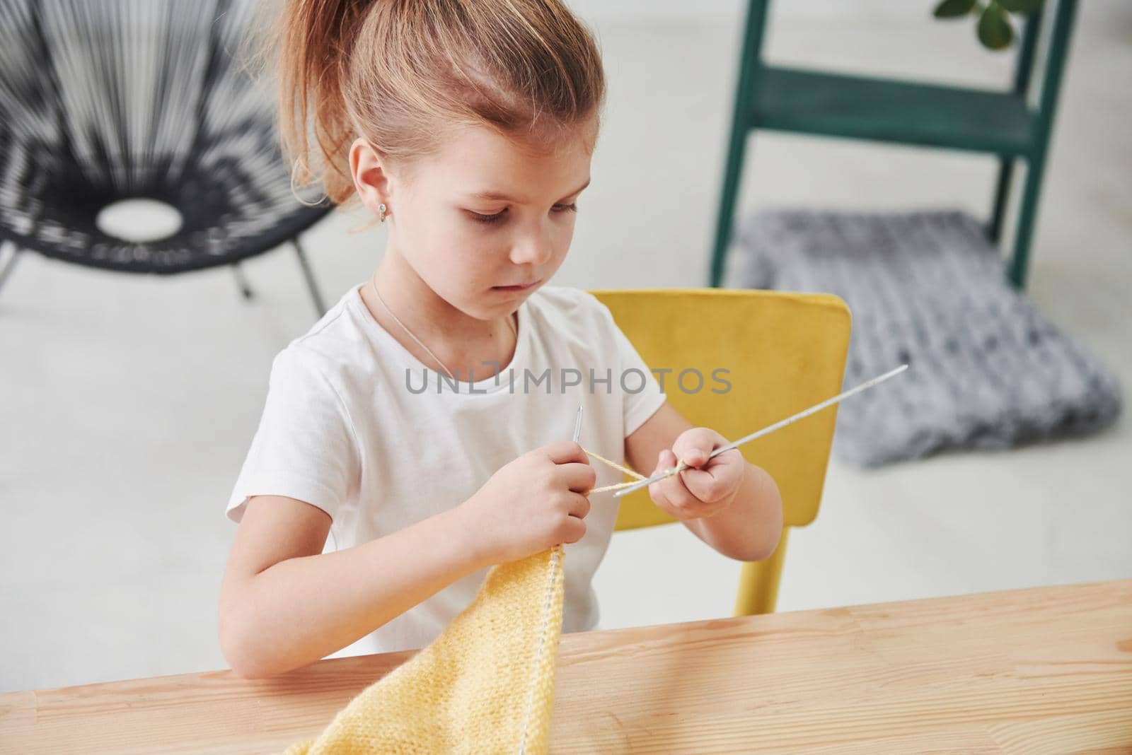 Trying her best. Kid is knitting at home. Cute little girl sitting near the wooden table is learning some new stuff.