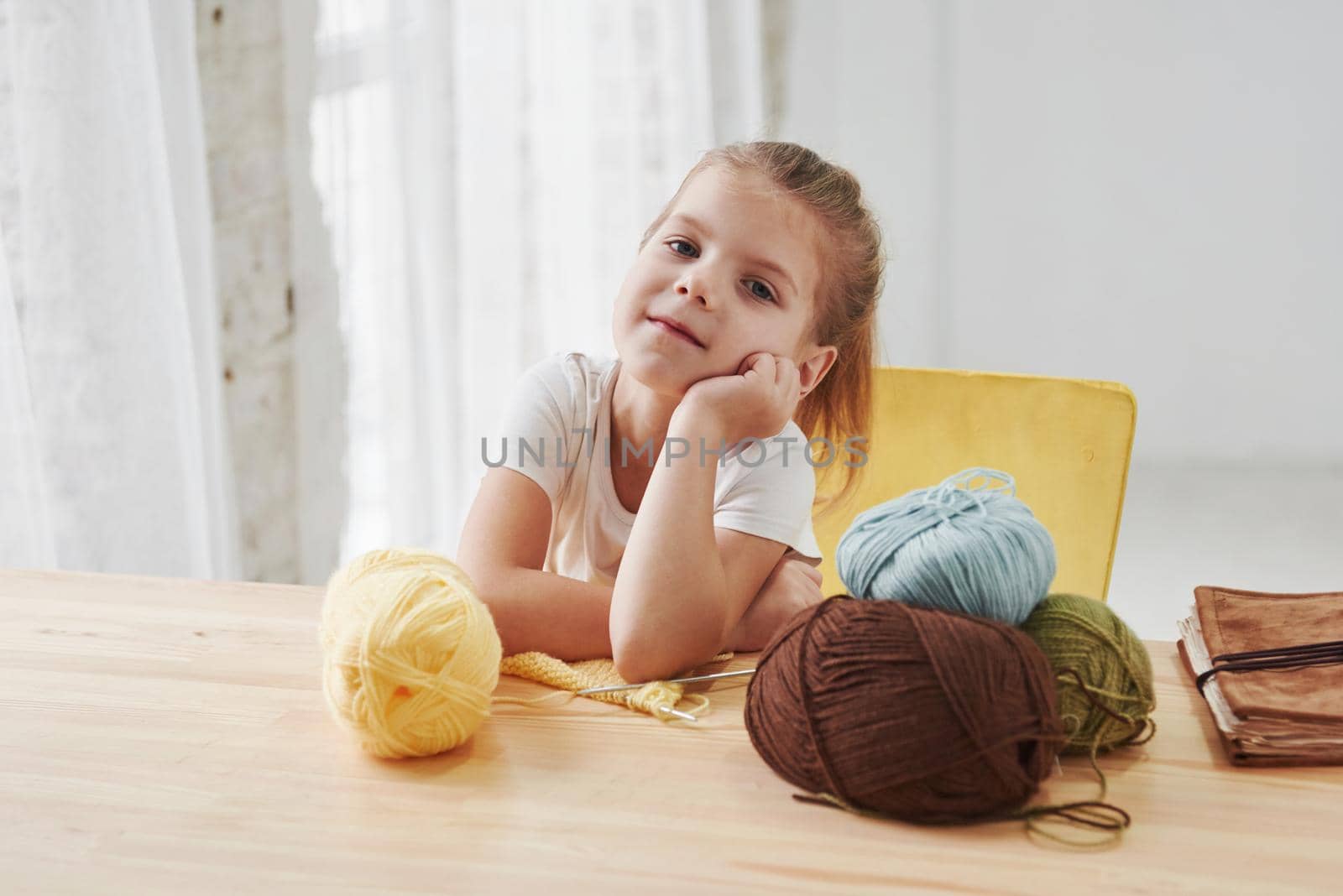 Quiet and calm interior. Kid is knitting at home. Cute little girl sitting near the wooden table is learning some new stuff.