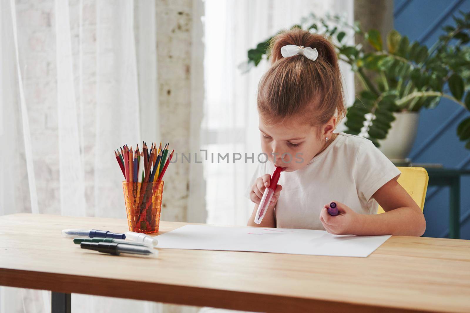 Holding marker in mouth. Cute little girl in art school draws her first paintings.