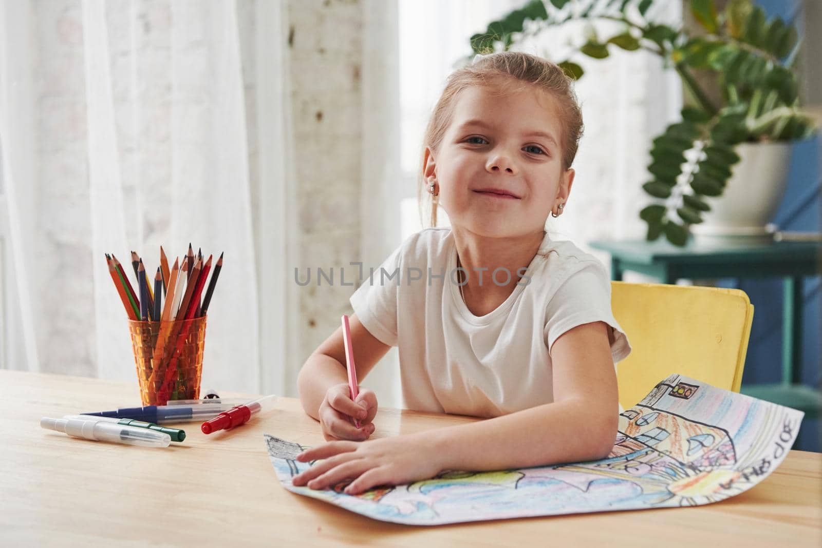 Looking into the camera with satisfied look. Cute little girl in art school draws her first paintings by pencils and markers.