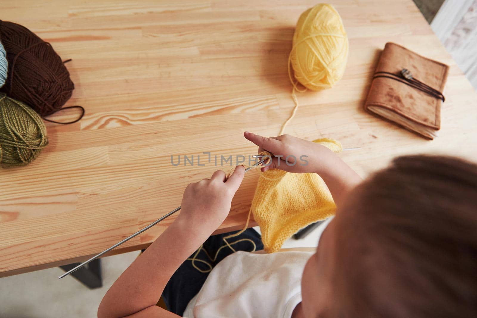 Close up view. Kid is knitting at home. Cute little girl sitting near the wooden table is learning some new stuff.