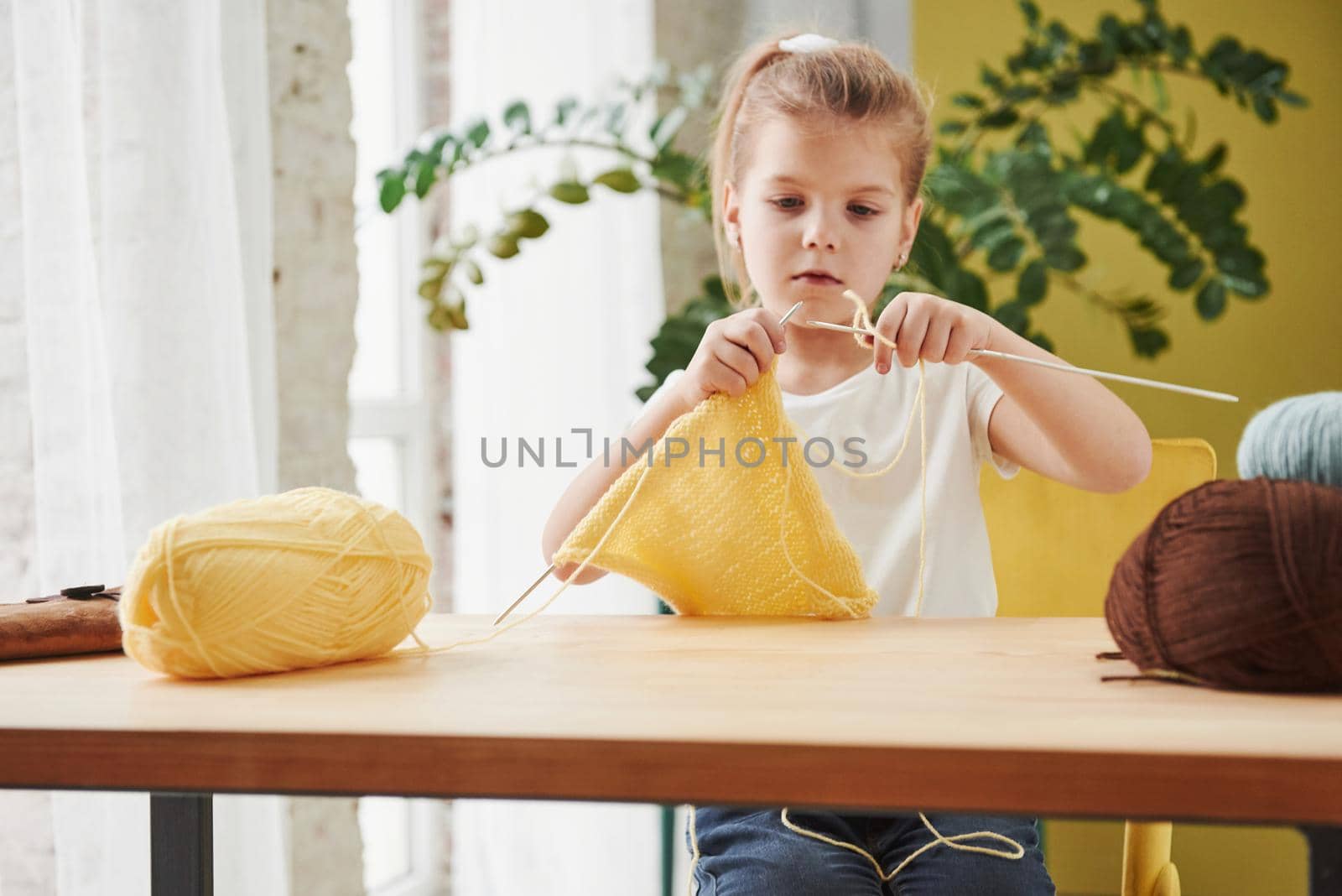 Looks like someone will have new cap. Kid is knitting at home. Cute little girl sitting near the wooden table is learning some new stuff.