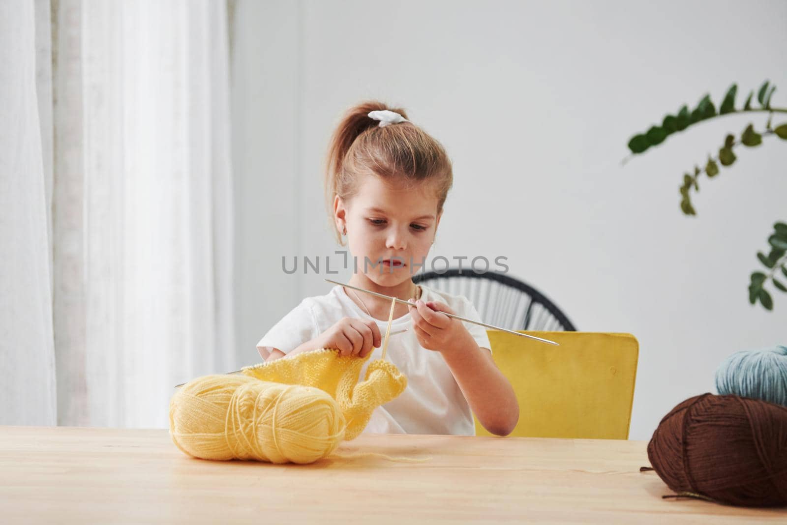 Yellow, brown and blue threads. Kid is knitting at home. Cute little girl sitting near the wooden table is learning some new stuff.
