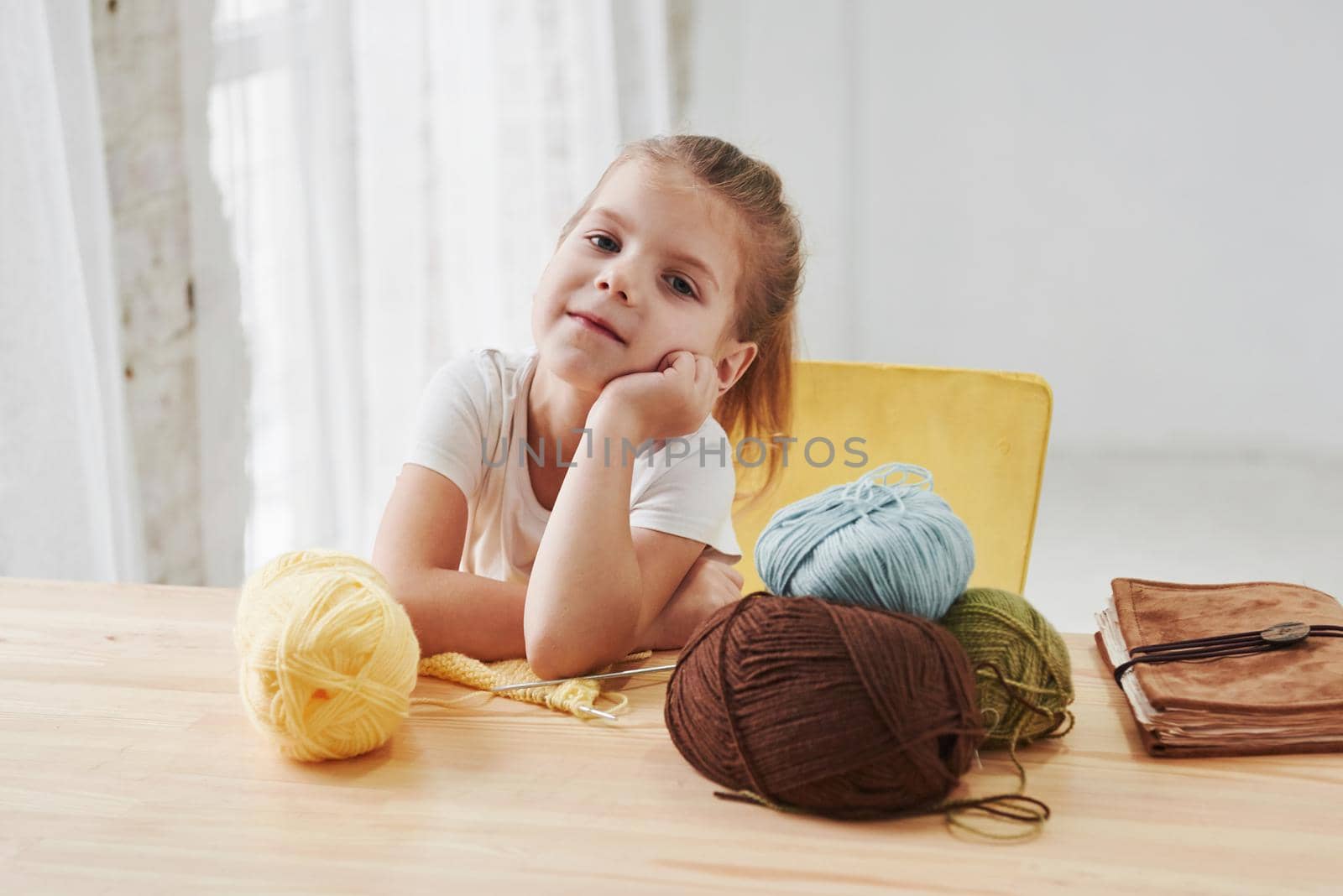 Posing for the portrait. Kid is knitting at home. Cute little girl sitting near the wooden table is learning some new stuff.