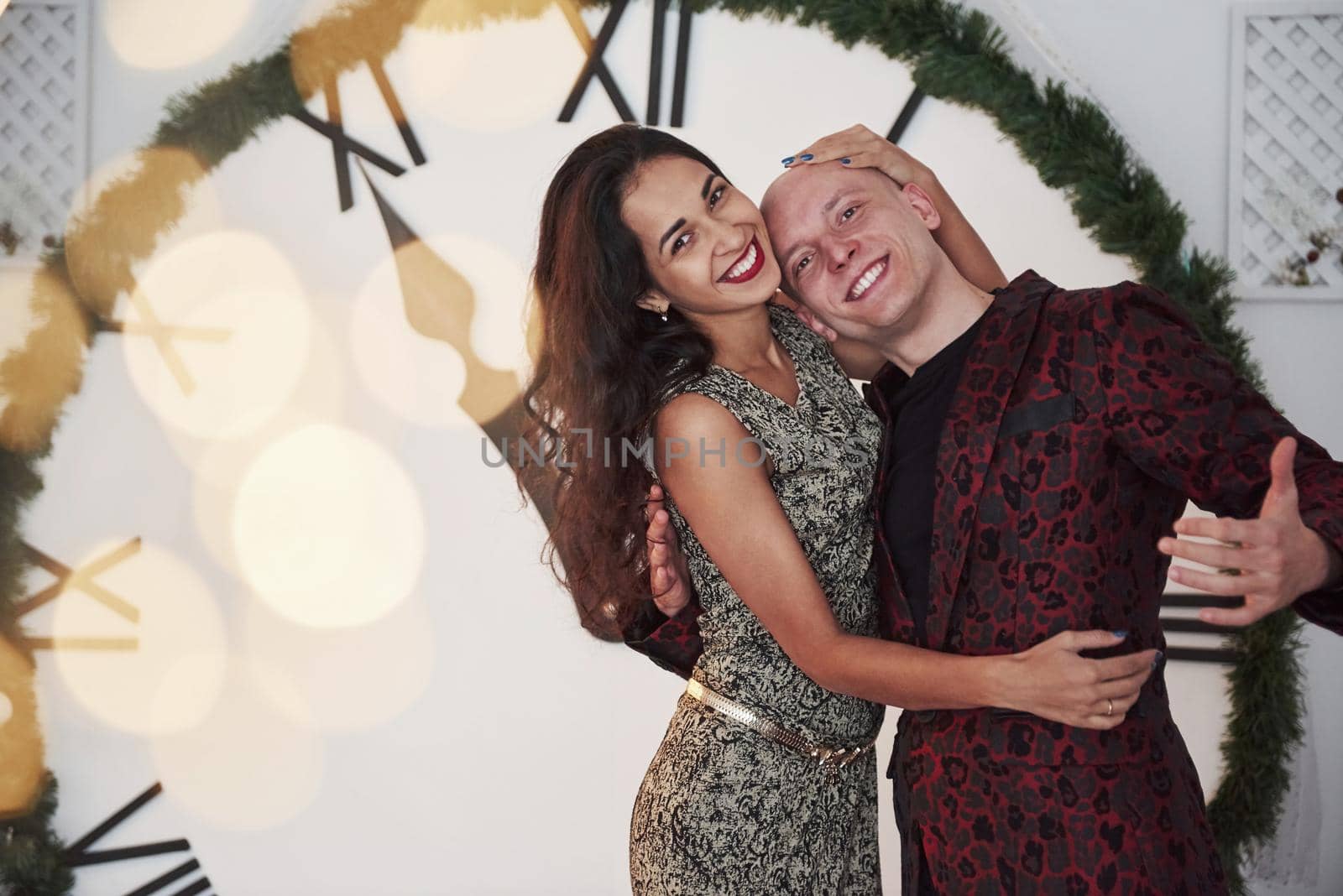 Cheerful mood. Couple in the holiday clothes. Big watch at background. Five minutes to New year.