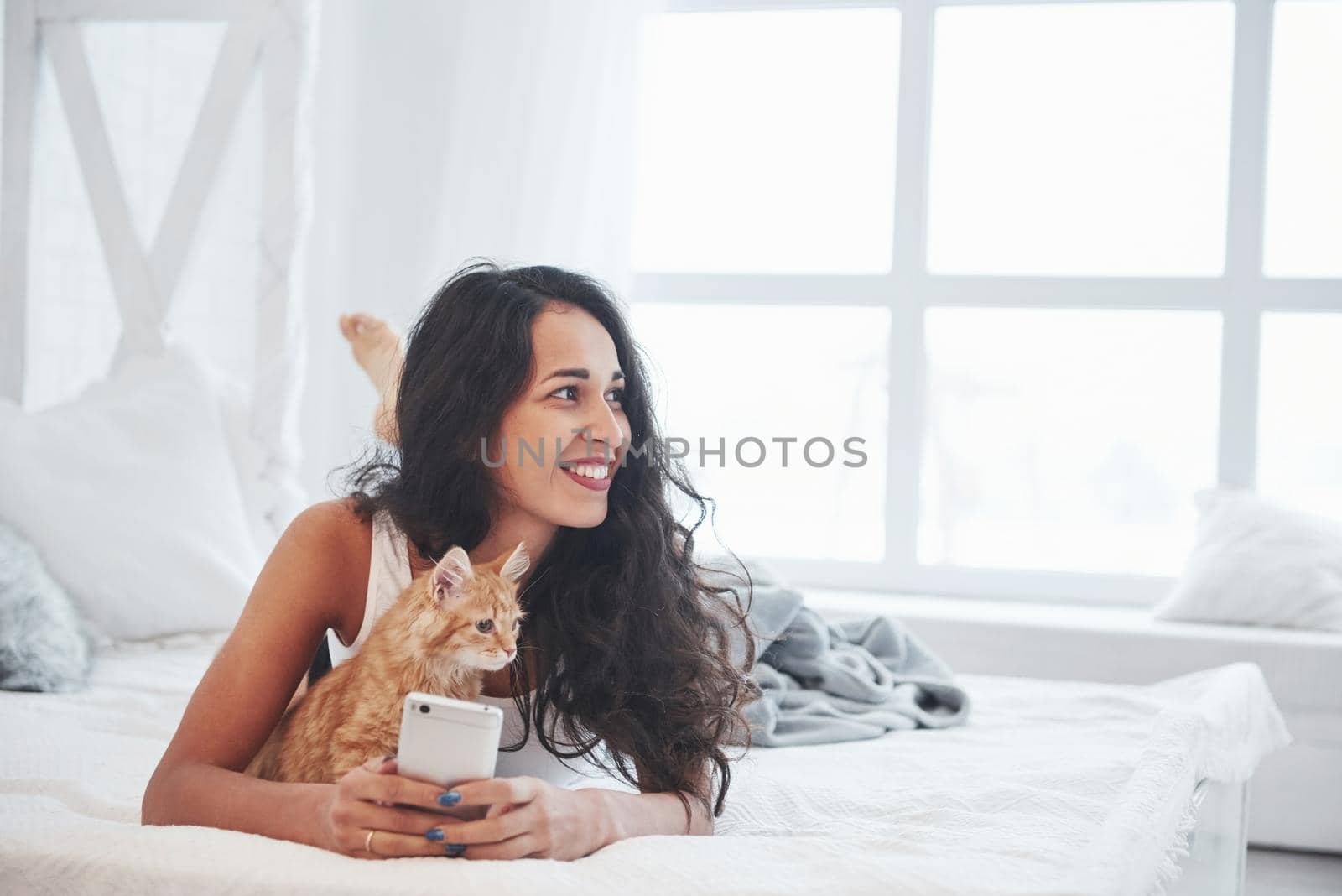 Looking to the side. Attractive blonde resting on the white bed with her cute kitten.