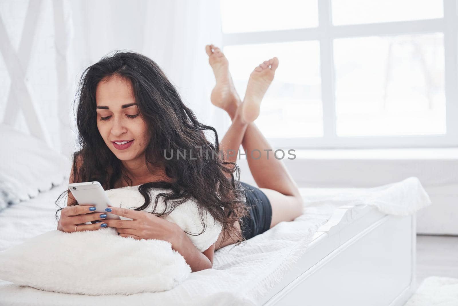 Conversation with friends using mobile device. Brunette having a rest on the white couch in her bedroom at daytime.