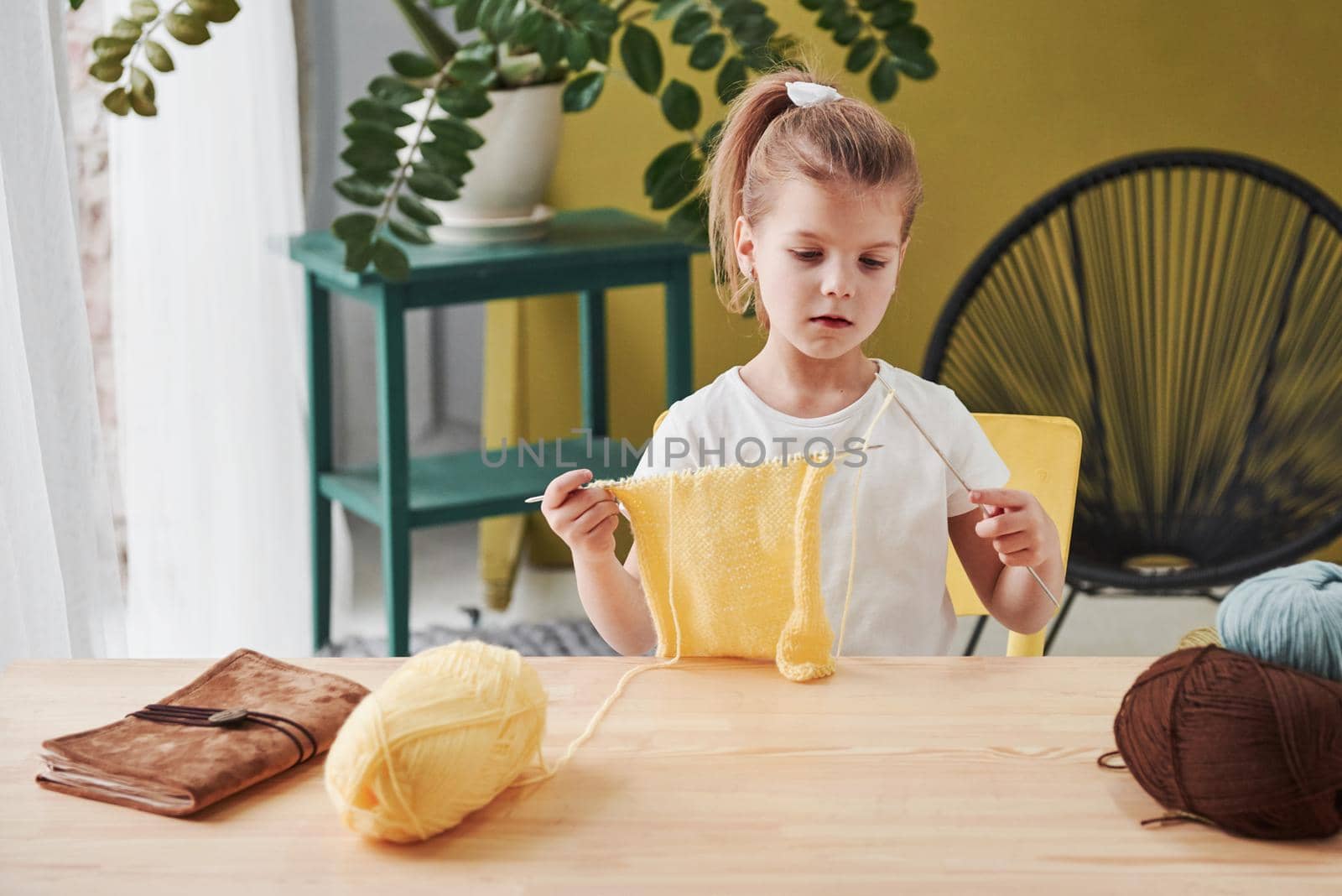 Concentrated at process. Kid is knitting at home. Cute little girl sitting near the wooden table is learning some new stuff.