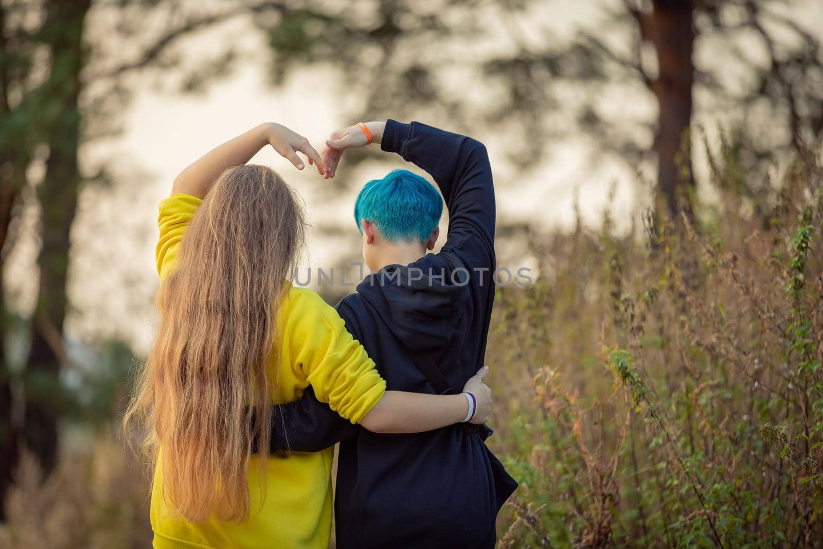 Two girlfriends are walking through the forest, raising their hands above their heads in the form of a heart. Girl with blue hair.