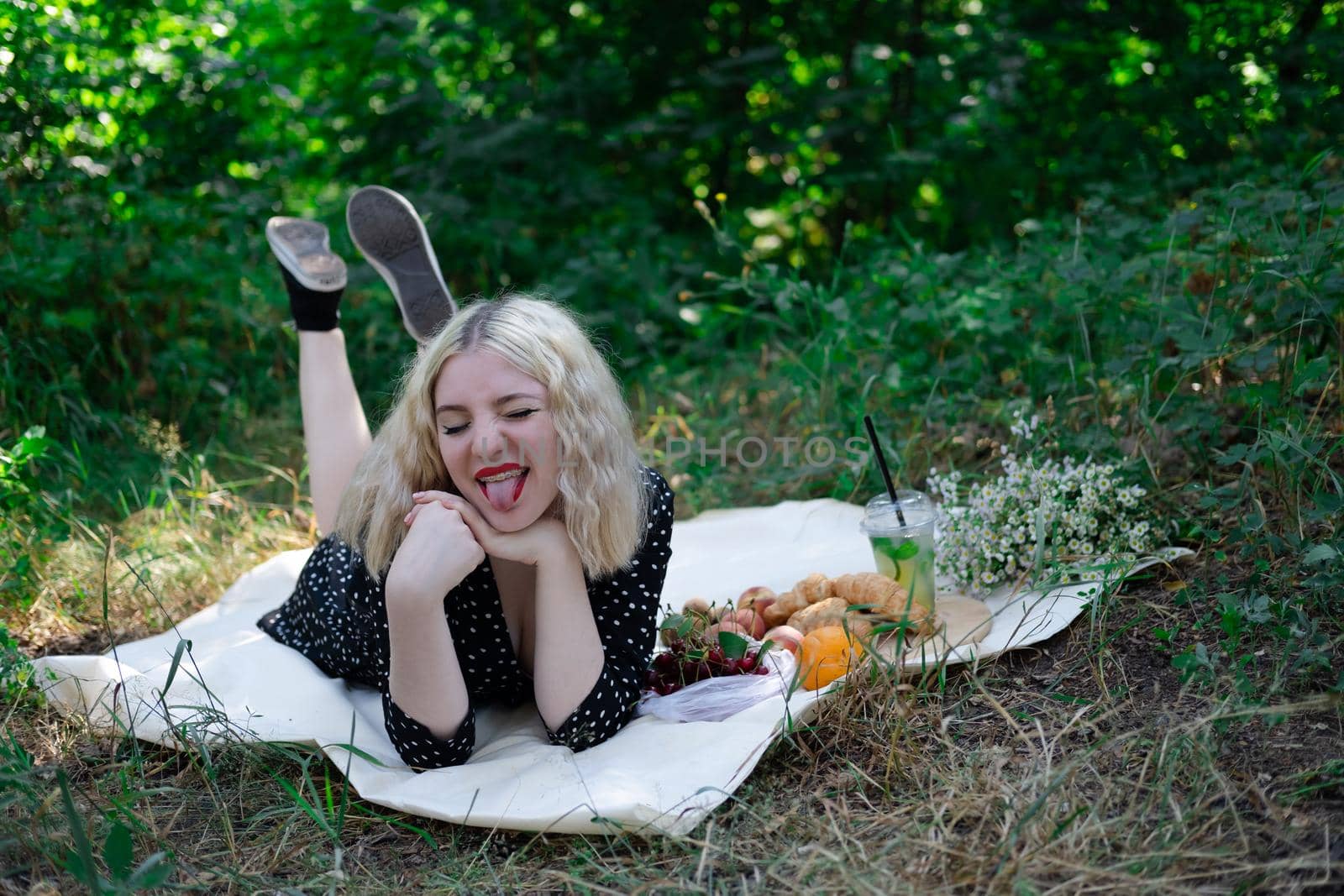 sweet blonde girl on picnic lying on white blanket in park. fresh summer refreshing drink and snacks. fruits and flowers.