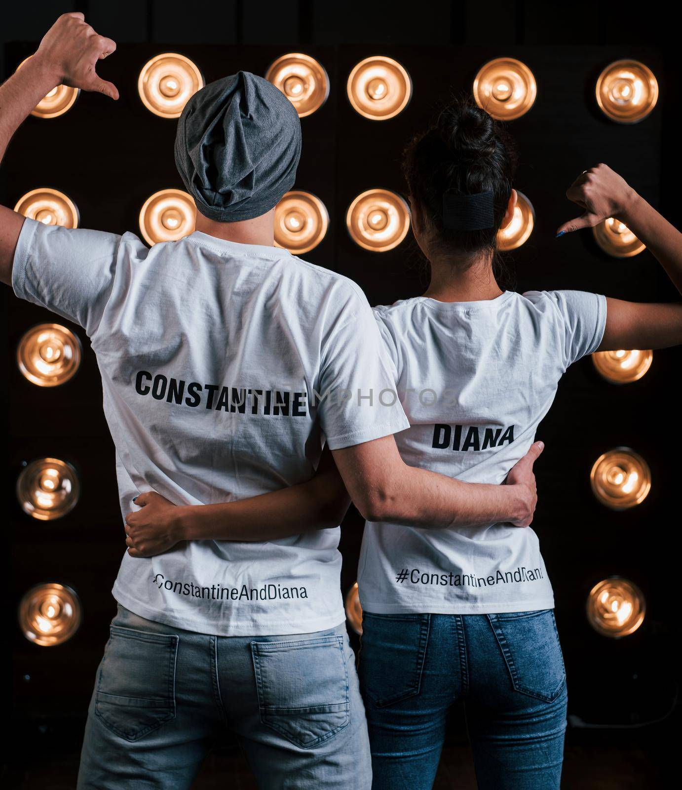 Shows thumbs on themselves. Back view of couple in shirts with their names on it standing next to studio lights.