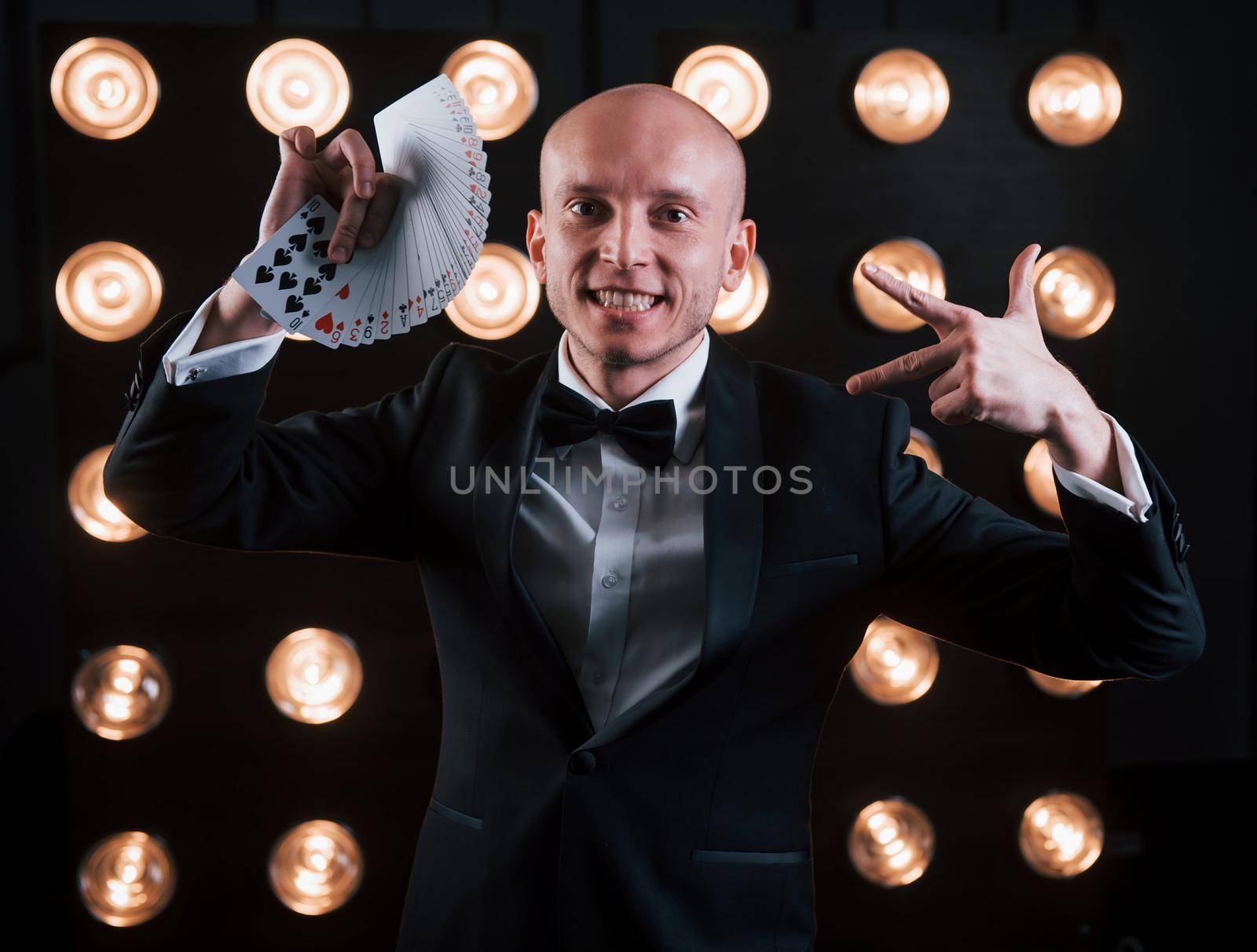 Showing some gestures. Magician in black suit and with playing cards standing in the room with special lighting at backstage.