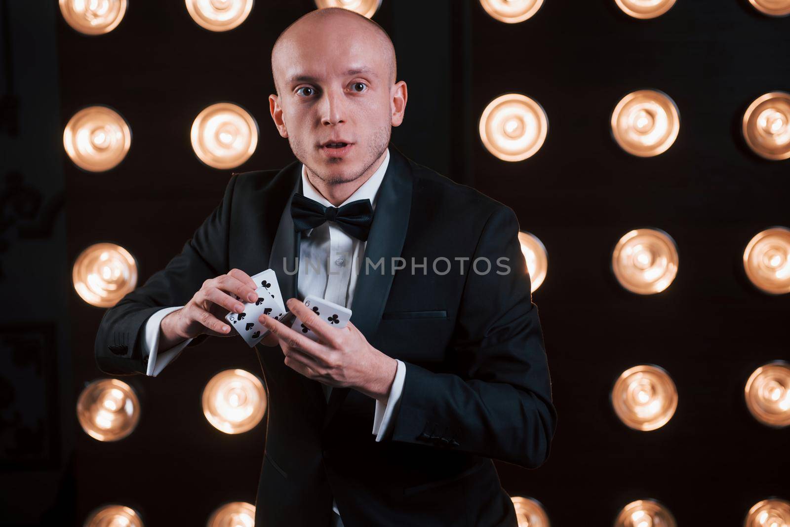 Shuffles the cards. Magician in black suit standing in the room with special lighting at backstage.