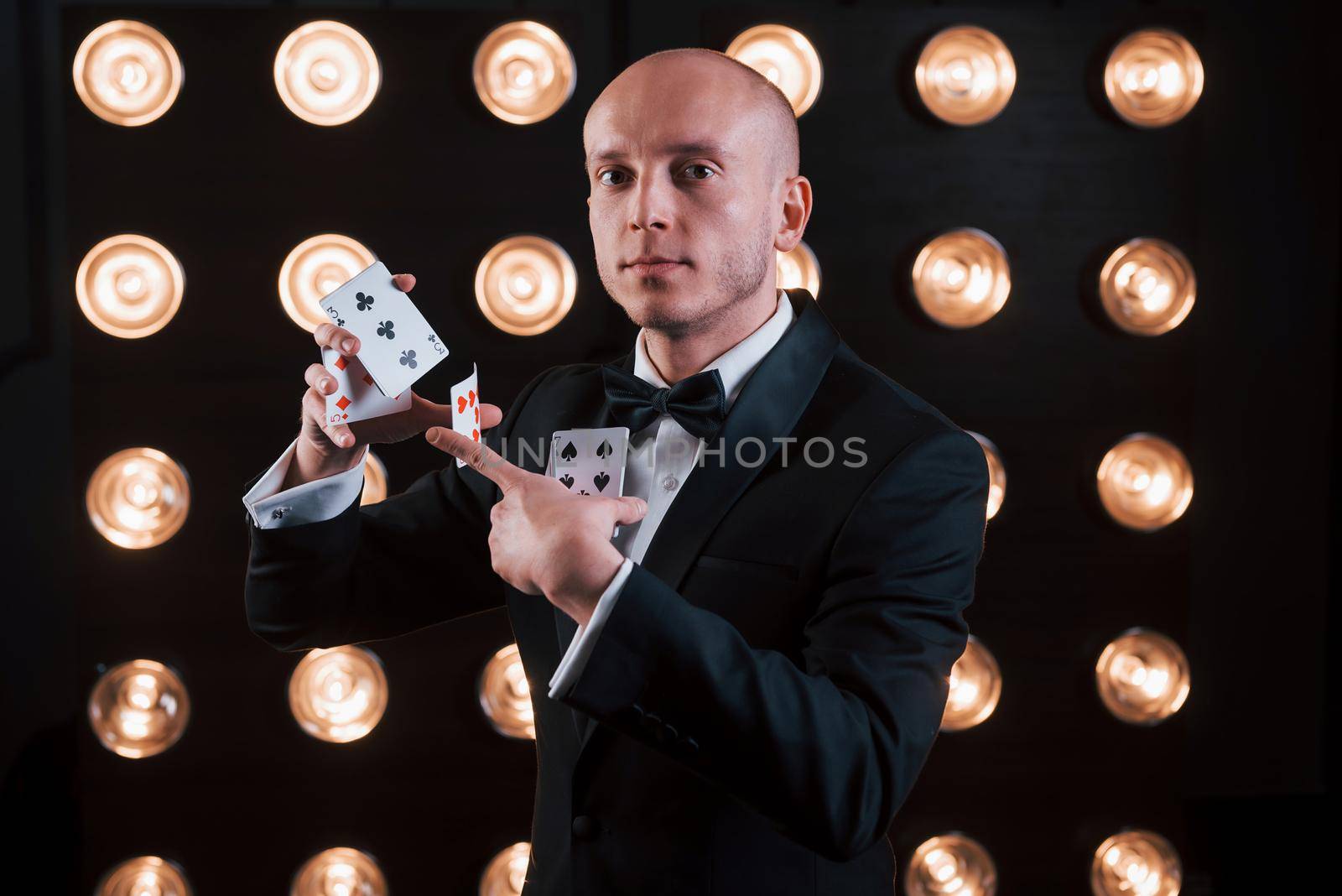 Some serious tricks. Magician in black suit and with playing cards standing in the room with special lighting at backstage.
