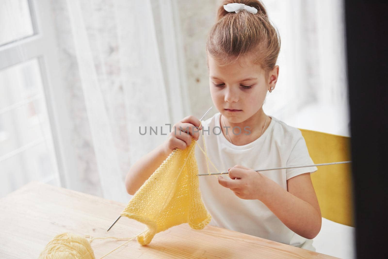 Daylight coming from the window. Kid is knitting at home. Cute little girl sitting near the wooden table is learning some new stuff.