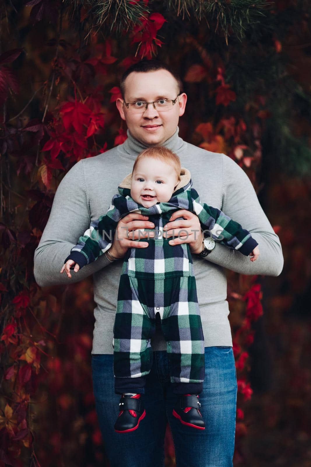 Professional portrait of attractive young man embracing his little baby girl in plaid warm overall while standing in beautiful bright tree with red leaves. They are smiling at camera surrounded by vivid foliage in autumnal park.