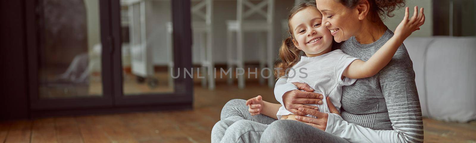 Jolly mother is hugging with little girl while doing workout together in cozy living room