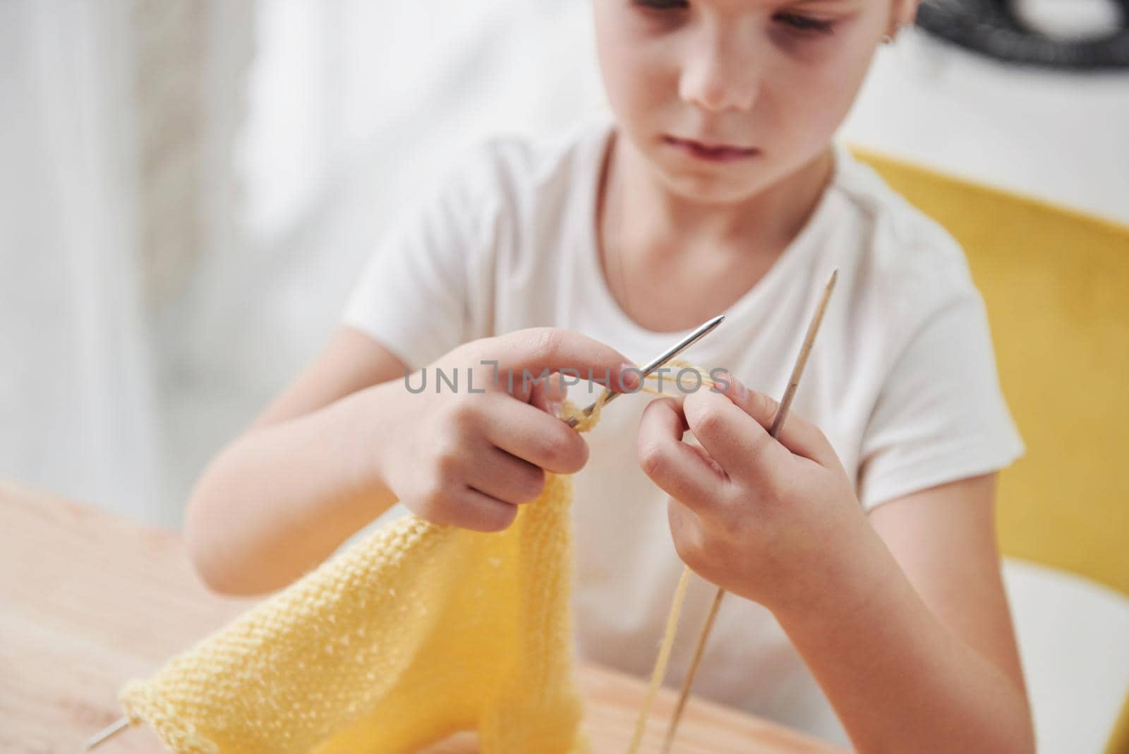 Close up view. Kid is knitting at home. Cute little girl sitting near the wooden table is learning some new stuff.