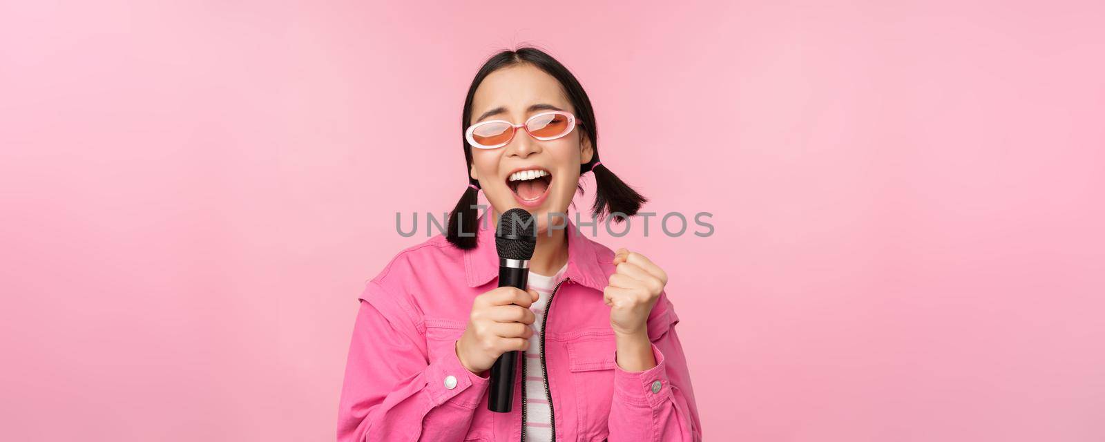 Happy beautiful asian girl singing with mic, using microphone, enjoying karaoke, posing against pink studio background.