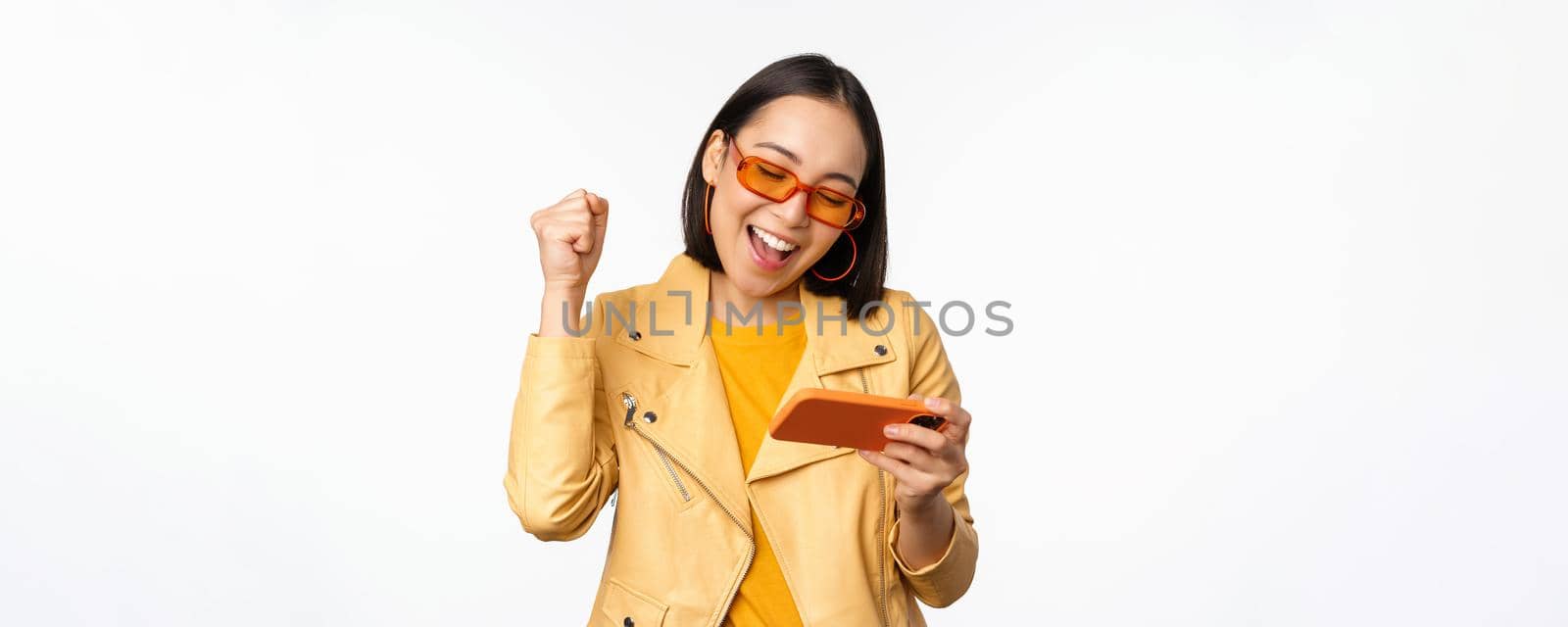 Stylish korean girl in sunglasses, playing mobile video game, laughing and smiling while using smartphone, standing over white background.