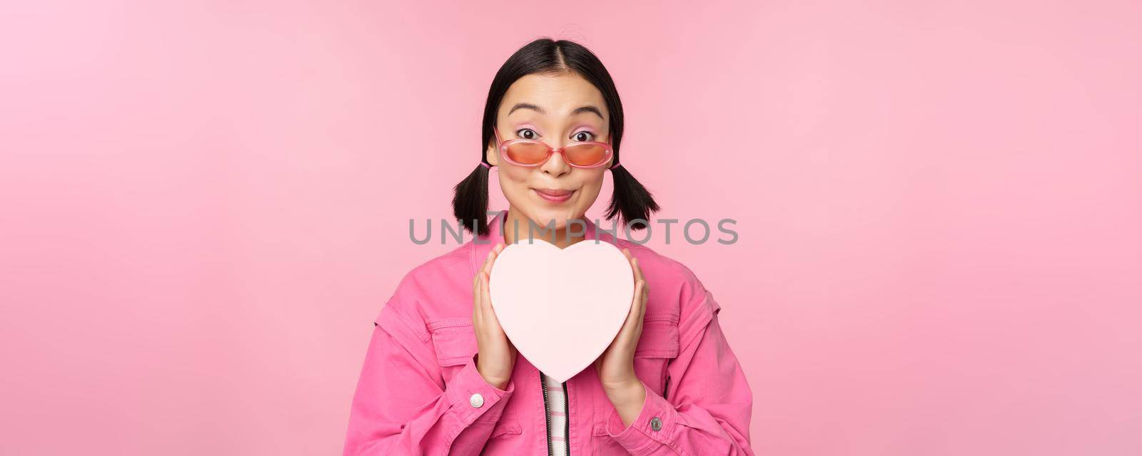 Beautiful asian girl smiling happy, showing heart gift box and looking excited at camera, standing over pink romantic background.
