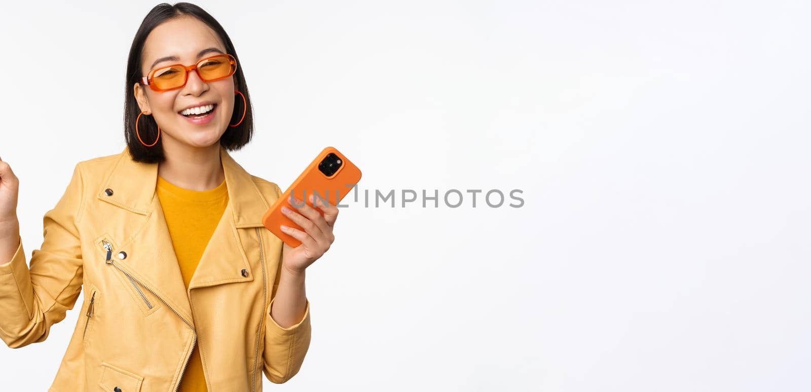 Image of stylish korean girl dancing with smartphone, laughing happy and smiling, standing over white background by Benzoix
